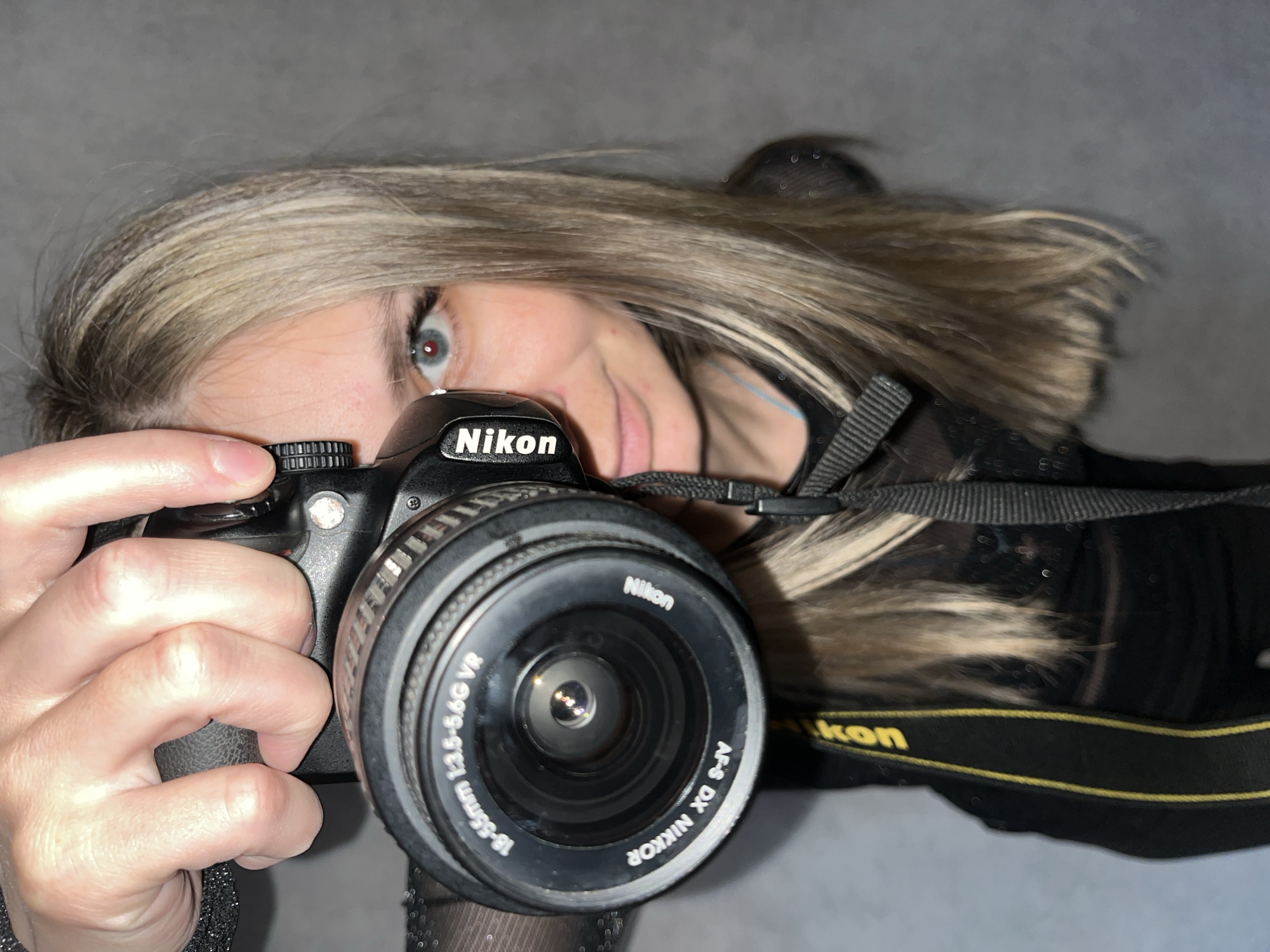 An individual with blonde and light brown hair is prominently featured in a close-up, angled shot, holding a black Nikon DSLR camera. Their right eye, a striking blue or grey, is visible, looking directly towards the viewer, suggesting a self-portrait or mirror shot. The person's left hand is wrapped around the camera grip, while their right index finger rests near the shutter button.
The camera is clearly identifiable as a Nikon model, with the brand name "Nikon" visible on the camera body and the lens. The lens attached is an "AF-S DX NIKKOR 18-55mm 1:3.5-5.6G VR," with this text clearly legible around its barrel. A black camera strap with yellow "Nikon" branding is slung around the person's neck, partially visible on the right side of the frame.
The background is a plain, undifferentiated grey, suggesting an indoor setting with minimal distractions. The lighting appears soft, illuminating the subject's face and the camera details. The person's expression seems focused, perhaps slightly curious or engaged, with a hint of a smile on the partially obscured mouth. The overall impression is a casual, direct capture of someone interacting with their photographic equipment. This image is set in Chaikovsky, Russia.