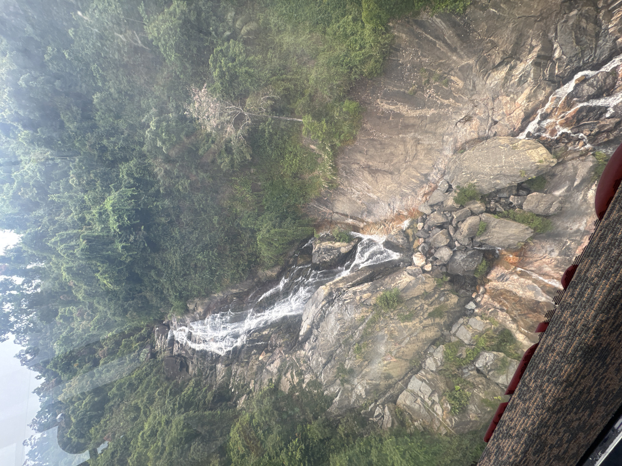 The image displays a majestic waterfall cascading down a rugged, rocky mountainside, viewed from an elevated perspective. The scene is dominated by a multi-tiered waterfall, with white, foamy water tumbling over dark grey and brown rock formations. Dense, verdant green forest covers the mountain slopes surrounding the waterfall, showcasing a lush, tropical environment. Some lighter-colored, possibly dead or dry, tree branches are visible among the green canopy.
The viewpoint appears to be from within a structure, likely a cable car or an observation deck, as indicated by the textured, dark-brown surface with red accents visible on the right side of the frame. This foreground element suggests the viewer is looking out through a window.
The time of day appears to be daytime, possibly under an overcast sky or slightly hazy conditions, which gives the scene a soft, diffuse light, particularly noticeable in the upper left portion of the image. No people or visible text are present, focusing the viewer's attention entirely on the natural landscape. The overall impression is one of natural grandeur and tranquility, characteristic of mountainous regions such as those found in Đà Nẵng, Vietnam.