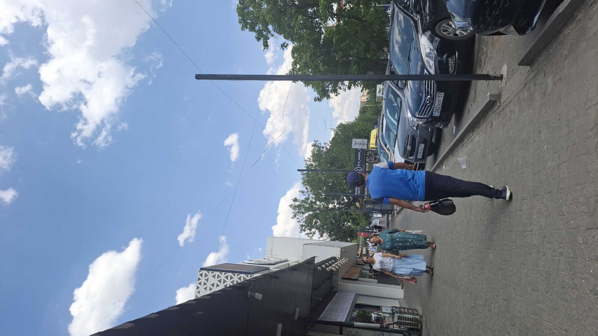 In Shymkent, Kazakhstan, a street scene unfolds under a partly cloudy sky. A man wearing a blue t-shirt and dark pants walks away from the viewer, carrying a plastic bag and a bottle. He walks on a paved sidewalk next to a row of parked cars, which includes a dark Hyundai with a license plate visible as "282 AUF 17". Two women walk on the sidewalk in the opposite direction. The woman closer to the viewer wears a white t-shirt and a light blue skirt, holding a red plastic bag. The woman beside her wears a green patterned dress. Shops with awnings are visible on the left side of the street. Trees line the right side of the street behind the parked cars. Power lines crisscross the sky.