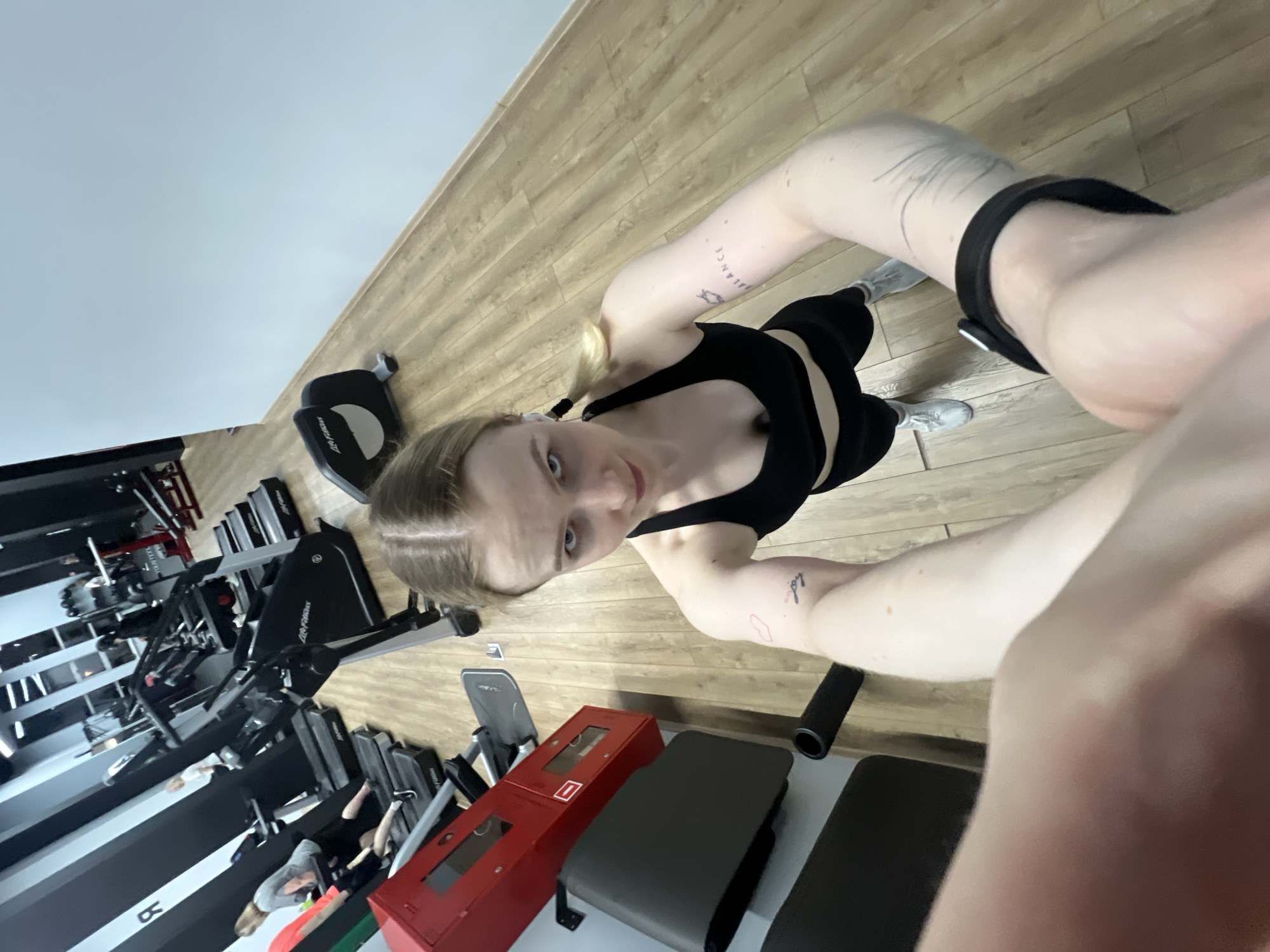 The image is a selfie taken by a young woman in a gym. She is wearing a black sports bra and black leggings. Her hair is blonde and pulled back in a ponytail. She has tattoos on her arms. In the background, there are treadmills and weight machines. There is a fire-resistant container next to some padded benches. From the available evidence, it is possible this is taken at a gym in Khimki, Russia.