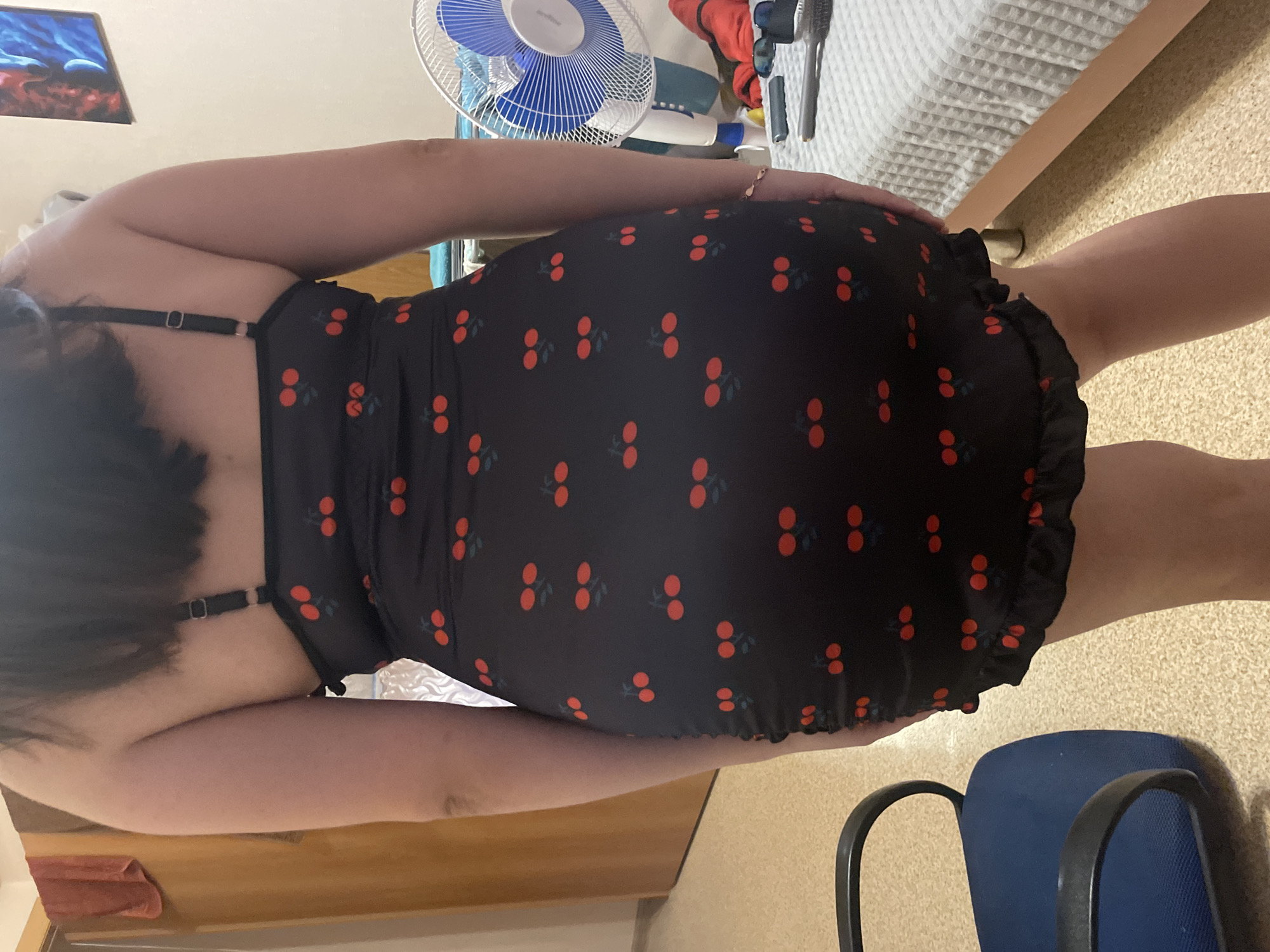 The image shows the back of a person from the waist down, wearing a black dress with a cherry pattern. The person is standing in a room with beige carpet and a blue office chair to the left. In the background, there is a white oscillating fan and a bed with gray bedding. A painting is visible on the wall above the fan. The room appears to be a bedroom or living space. There is no indication of the city of Laishevo, Russia within the image or its context.