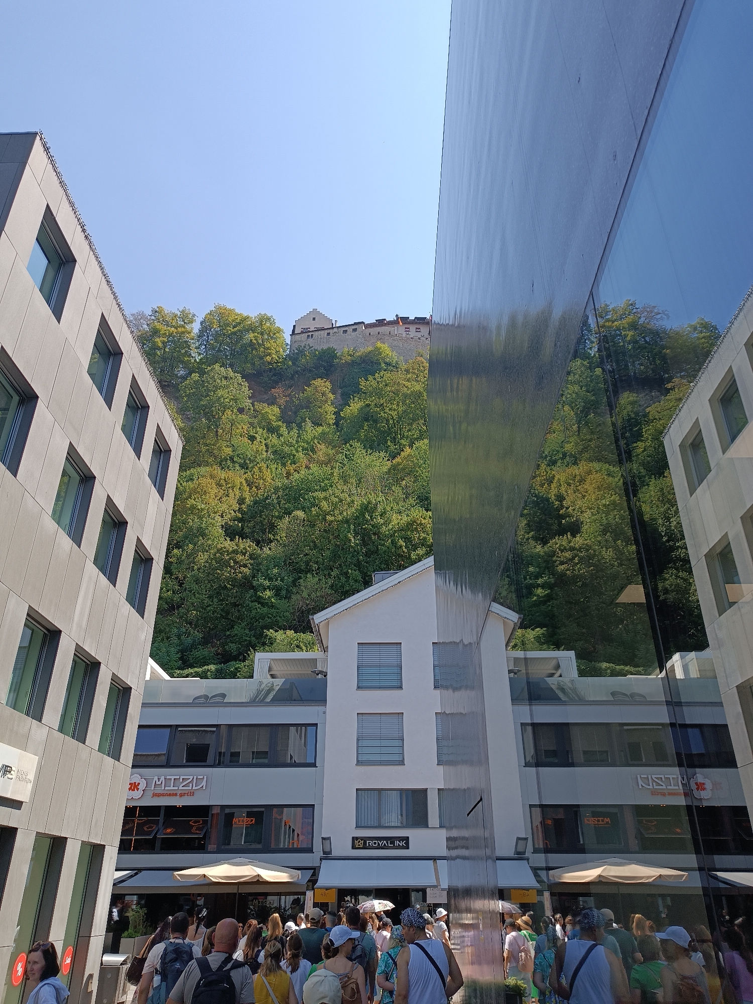 The image depicts a street scene in Vaduz, Liechtenstein. In the foreground, a crowd of people walks along a street lined with modern buildings. To the left, a contemporary building with a gray, textured facade and numerous windows is visible. To the right, a highly reflective, dark glass facade of another modern building dominates the view, mirroring the surrounding environment, including trees and a castle on a hill. In the midground, a white building with multiple windows is situated between the two modern structures. Above the buildings, a lush green hillside is covered with trees, and at the top of the hill sits a historic castle. The sky is clear and blue. The overall impression is of a sunny day in an urban environment with a prominent historical landmark.