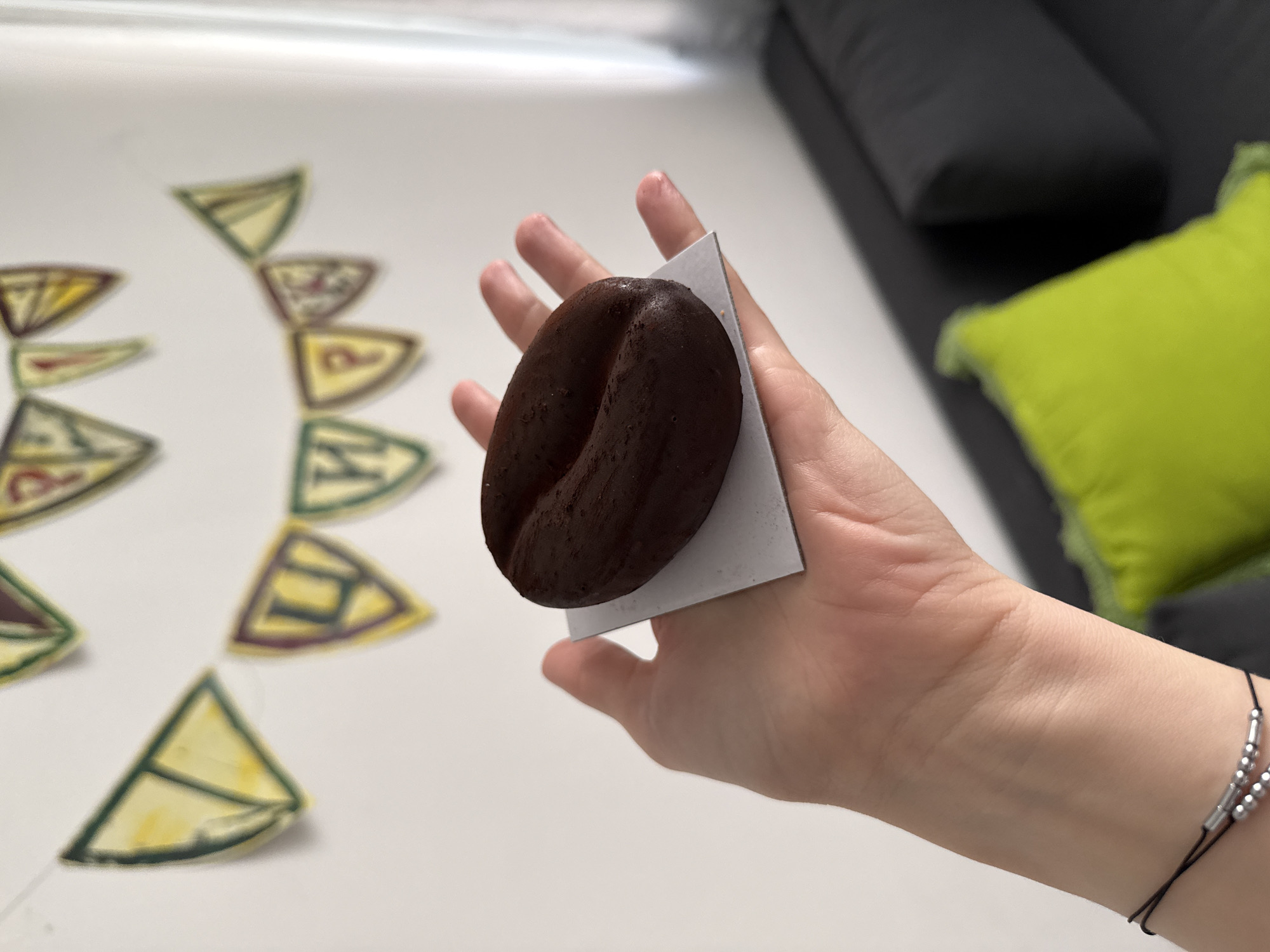 A human hand is holding a dark brown, kidney-shaped object (possibly a chocolate molded to resemble a coffee bean) atop a small, square, light gray or white piece of cardstock. The hand has a bracelet consisting of silver beads on a black cord around the wrist. In the blurred background, there are multiple small, triangular pieces of paper with drawings on them arranged in a semi-circle on a white surface. To the right, there is a section of a dark gray couch with a bright green square pillow. The location of Ramat Gan, Israel is not detectable from the image.