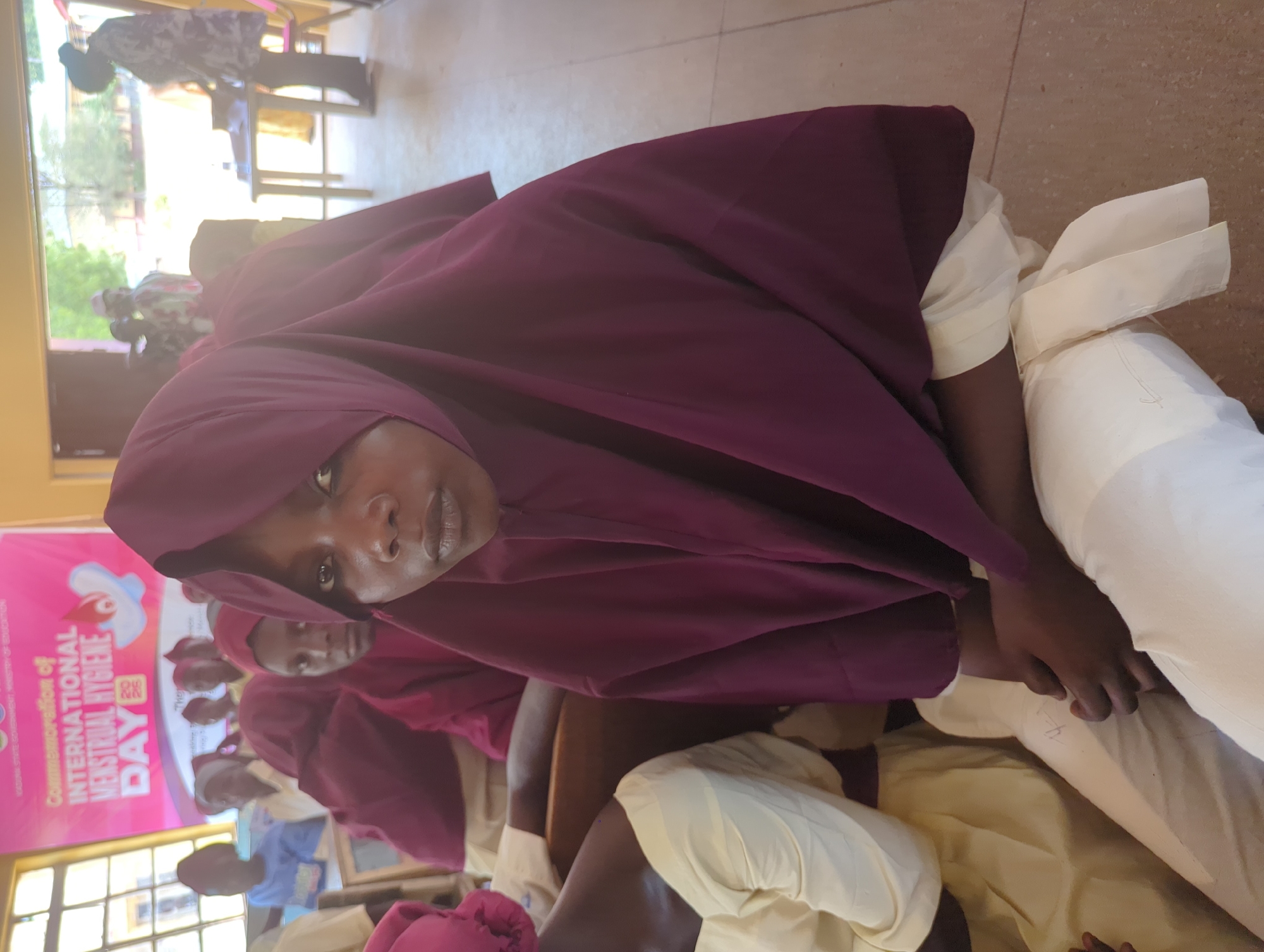 This media file depicts an indoor gathering, likely a school or community event, in Kakuri Gwari, Nigeria. The subjects are primarily young people, predominantly girls, dressed in modest clothing including hijabs and what appear to be school uniforms in light yellow and cream colors. The main subject in the foreground is looking directly at the camera with a neutral expression.
The event seems to be related to "International Menstrual Hygiene Day," as indicated by a large pink banner visible in the background. The banner displays text in English related to the commemoration of this day, including the year "2022". There are other individuals in the background, some seated and some standing, also appearing to be participants in the event. The setting suggests an educational or awareness-raising environment. The lighting appears to be artificial, suggesting an indoor space, and the time of day is not discernible. The atmosphere seems to be one of a structured gathering or presentation.