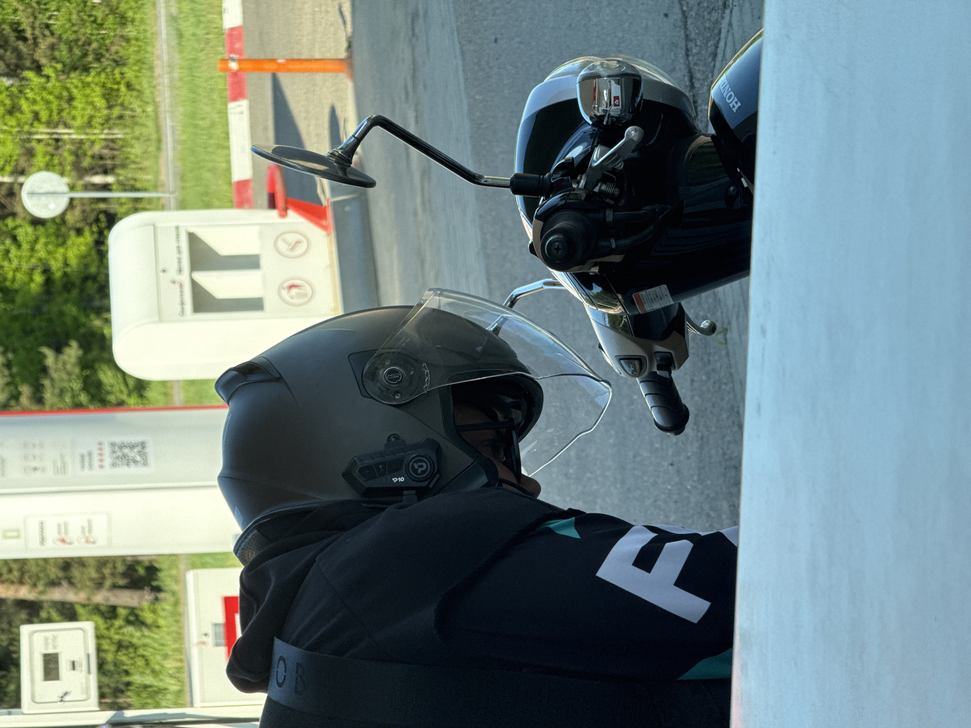 The image shows a person wearing a grey helmet with a clear visor, seated on a Honda motorcycle. The location appears to be a gas station, with a gas pump visible in the background. The background also shows greenery. The person is wearing a black jacket with white text. The motorcycle is black and silver. The photo appears to be taken in Chaikovsky, Russia.