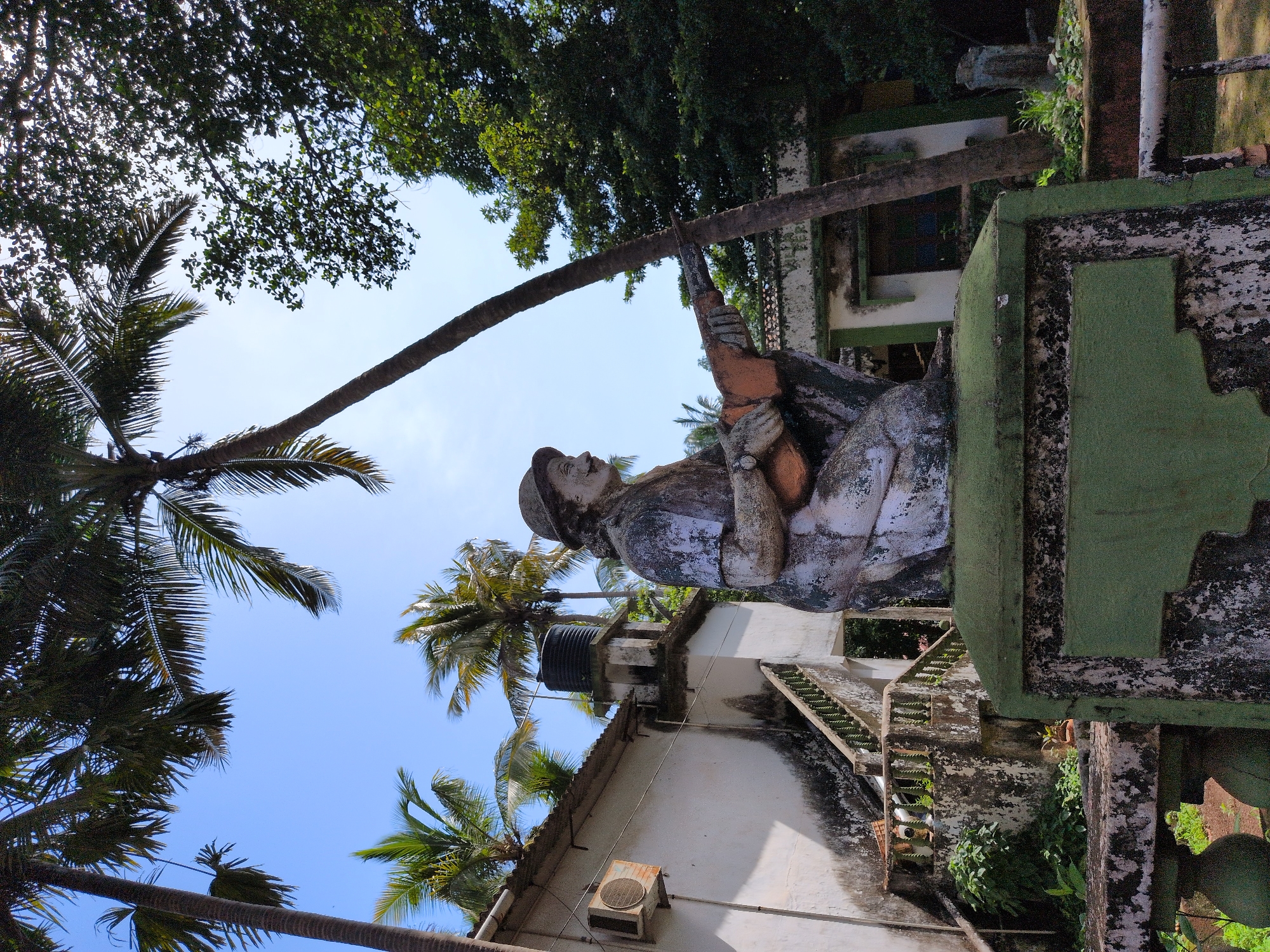 A statue of a soldier, possibly made of concrete or plaster, is positioned in the foreground. The statue is weathered, with peeling paint and moss growing on its surface. It depicts a figure in a helmet, kneeling and holding a rifle. Behind the statue, a weathered white building with an outdoor air conditioning unit is visible, along with a water tank on the roof. A concrete staircase with a green railing leads up to the building. Palm trees are prominent throughout the scene, with lush green foliage and clear blue sky overhead. The overall impression is of an abandoned or neglected outdoor space in a tropical or subtropical environment. The location is the war memorial in Oxel, India.