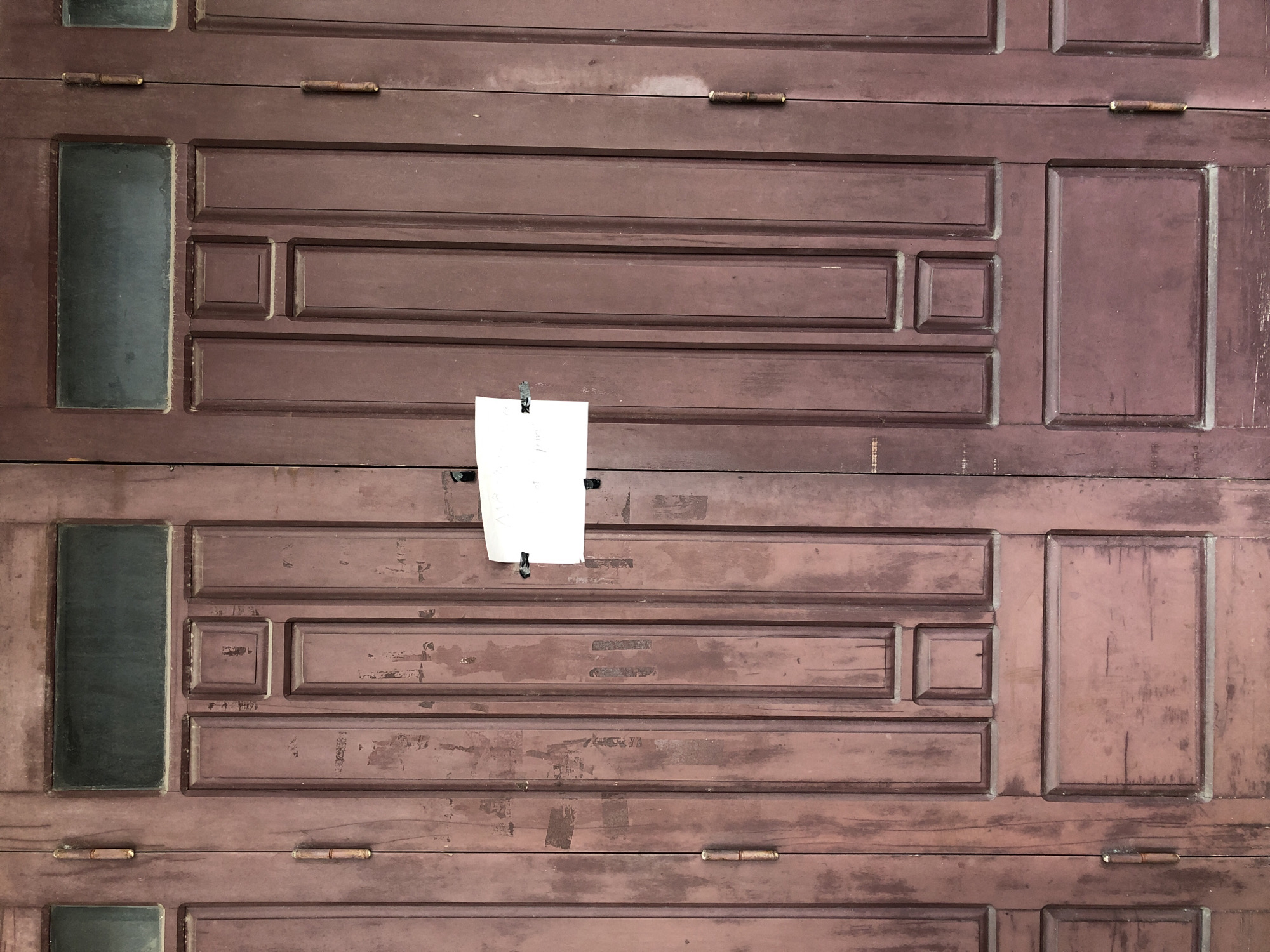 A pair of weathered, dark red wooden doors in Da Nang, Vietnam, are partially open, revealing a sliver of darkness within. The doors are divided into multiple panels, with decorative trim outlining each section. A small, white piece of paper is taped to the right-hand door, near the center. Two hinges are visible on each door, suggesting they are old and possibly heavy. The wood appears worn and faded in places, with some scuff marks and scratches. Above the panels, each door has a narrow, rectangular glass window. The overall impression is one of age and perhaps disuse.