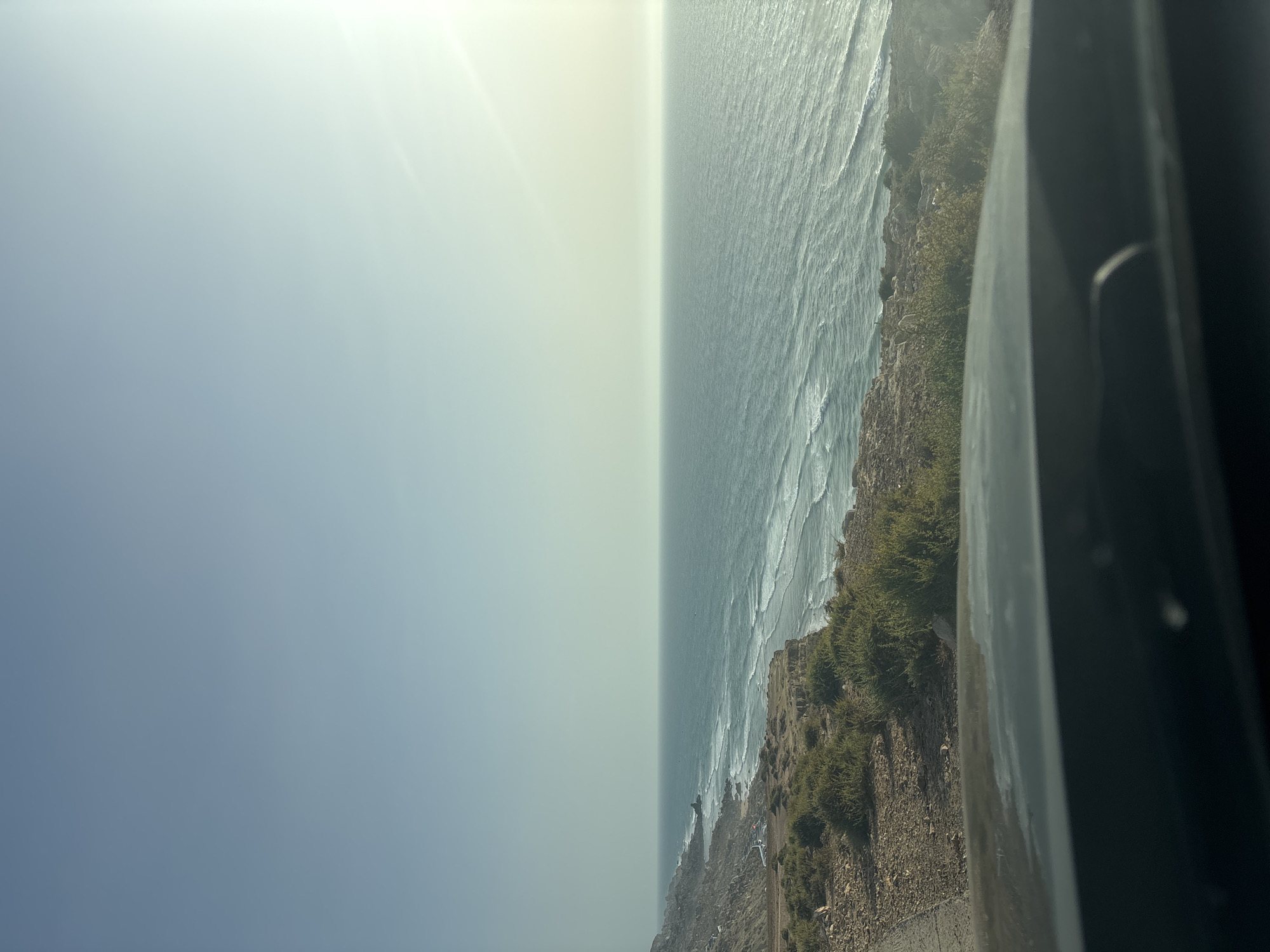 A wide-angle, elevated shot captures a serene coastal landscape under a hazy, bright sky. The foreground is dominated by the dark, reflective surface of a car's dashboard. Beyond this, a rugged, rocky coastline stretches out to the left, sparsely populated with green shrubs. A narrow, unpaved road runs alongside the cliff edge. The ocean, a vibrant turquoise, meets the shore with gentle, rolling waves. The horizon is a soft, pale line where the sea meets the pale, almost white sky, with the sun's glare creating a diffused bright spot on the right side.