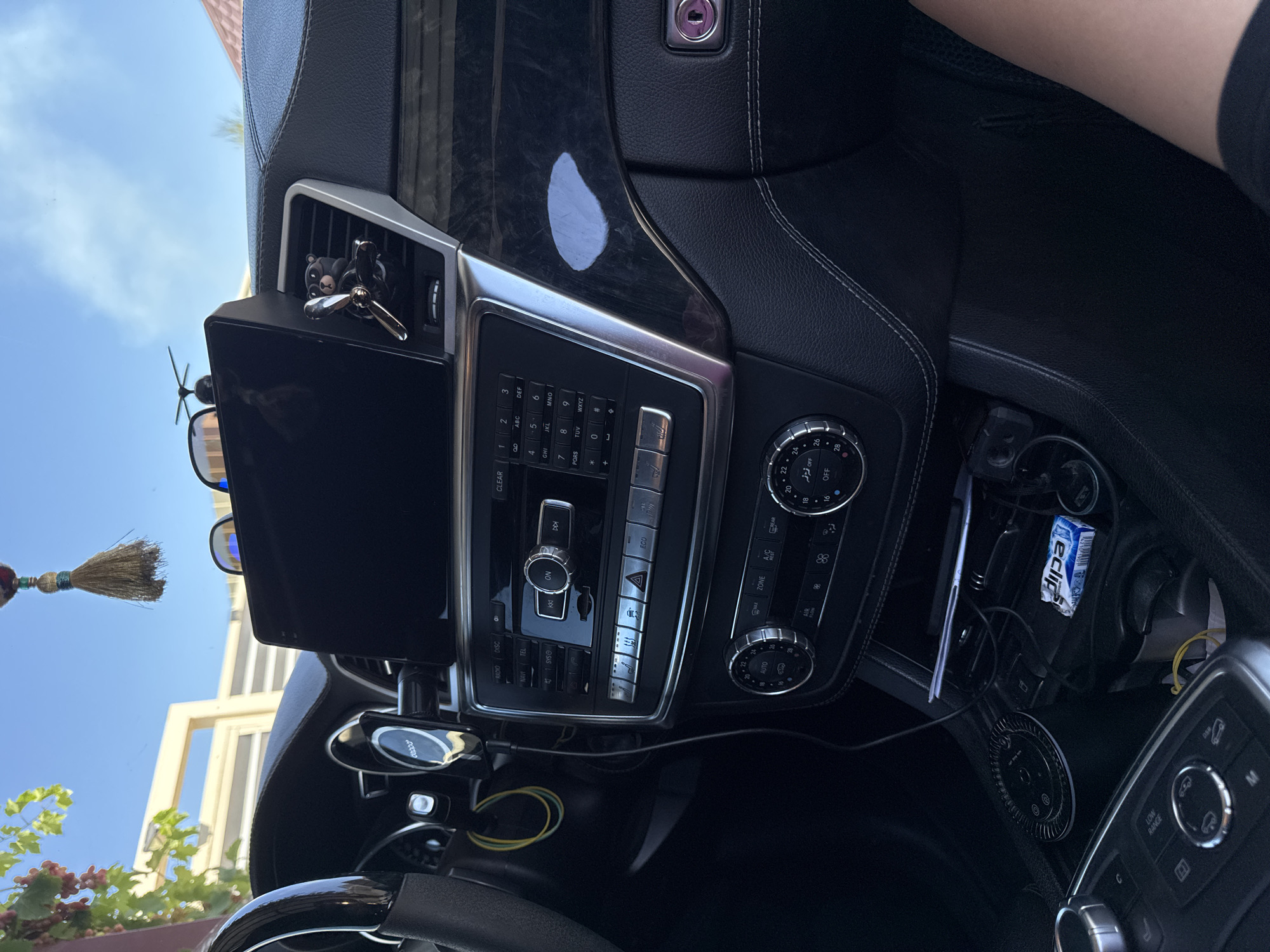 The image shows the interior of a car, specifically the dashboard and center console area. The car appears to be parked outdoors, with a bright sky visible through the windshield.
Key objects in the scene include:
- A large, black infotainment screen mounted above the dashboard.
- Various car controls, including buttons, knobs, and a dial for climate control.
- A small, decorative item resembling a bear with sunglasses and a propeller attached to one of the air vents.
- A mobile phone mounted on the left side of the screen.
- Charging cables and a pack of gum in the center console.
- A person's leg and knee are visible on the right side of the frame, suggesting someone is in the driver's or passenger's seat.
The overall impression is of a car interior with some personal items and decorations. The image's detail allows for a forensic examination of the car's interior features and the presence of personal belongings. The location is stated as Uyutnoye, Ukraine, providing a geographical context to the scene.