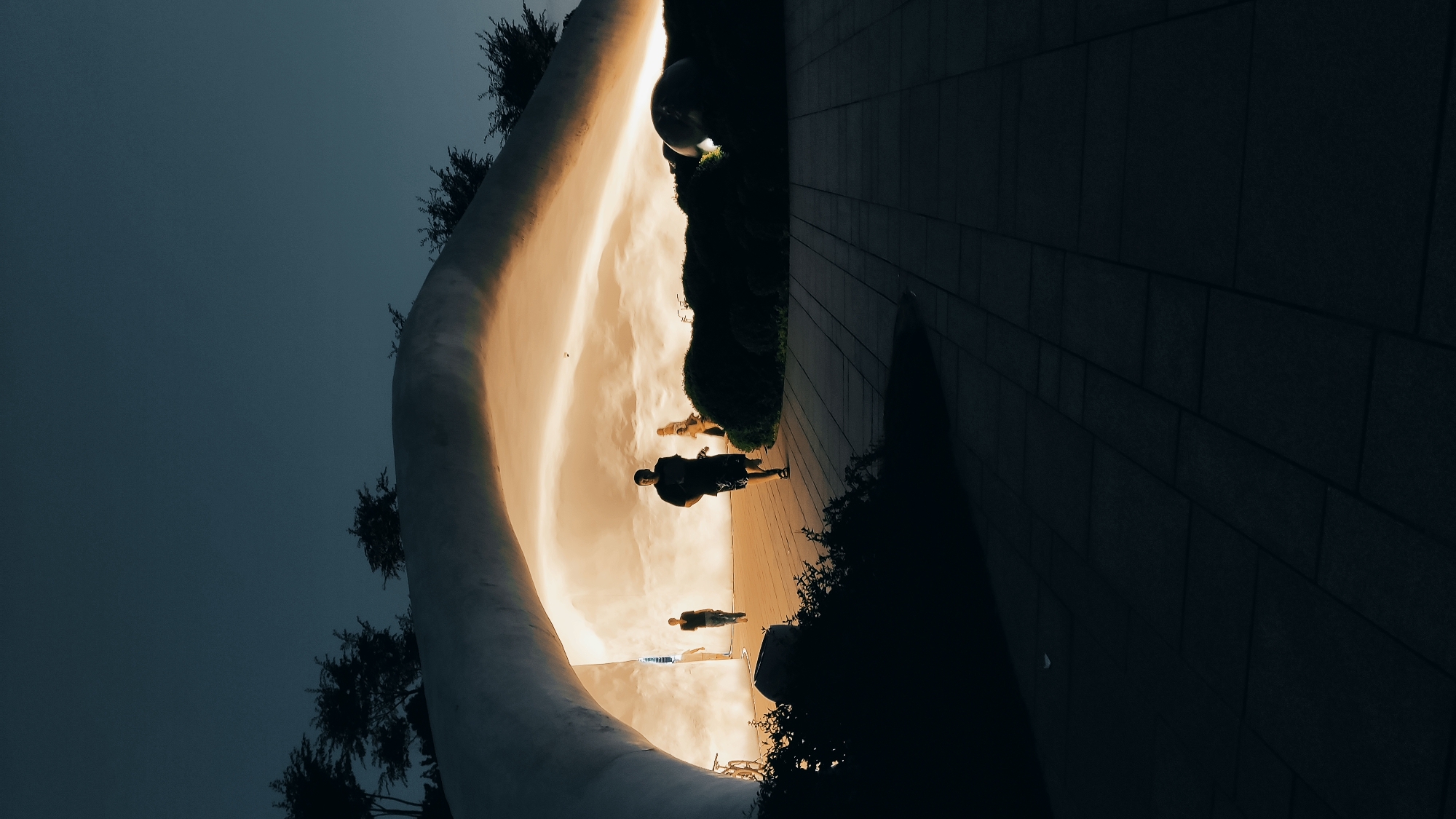 This image captures an outdoor scene at dusk or night, featuring a dramatic interplay of light and shadow. The primary visual element is a large, smooth, light-colored curved wall on the left, which is brightly illuminated with a warm, golden glow. This intense light creates a strong contrast with the dark, muted blue-grey sky above and the deeply shadowed, paved ground on the right, which shows a pattern of rectangular tiles.
Several individuals are present, appearing as dark silhouettes against the glowing backdrop of the curved wall. In the mid-ground, a person stands, their form clearly outlined by the light. Further back and to the left, two more human figures are visible as smaller, indistinct silhouettes, suggesting others in the space. Dark, dense bushes or foliage line the base of the curved wall and extend along the right side of the frame, partially obscuring a large, dark, reflective spherical object nestled within them in the mid-background.
The scene conveys a quiet and contemplative mood, enhanced by the low light and the anonymous, silhouetted figures. The sleek design of the curved wall and the paved pathway suggest a modern architectural or public park setting. The illumination of the wall provides the main light source, highlighting its form and casting the surrounding elements into deep shadow. There is no visible text in the image.
