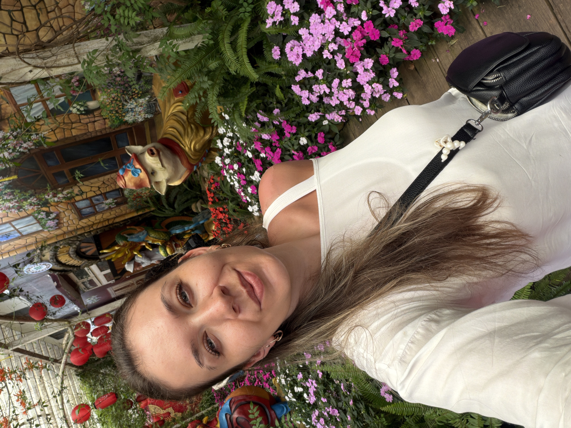 A selfie shows a young Caucasian woman with long brown hair and blue eyes smiling in front of a colorful garden display. She is wearing a white off-the-shoulder top and a black crossbody bag. The background features vibrant pink and white flowers, lush green ferns, and a whimsical stone building with ornate details. Red lanterns are suspended from the ceiling, adding to the festive atmosphere. In the middle ground, a golden, anthropomorphic pig statue wearing a hat and red scarf is visible amongst the greenery. The woman is positioned slightly to the left of the frame, with the garden filling the rest of the image. This scene is likely located at the Datanla New City in Da Lat, Vietnam, known for its creative garden displays and themed attractions.