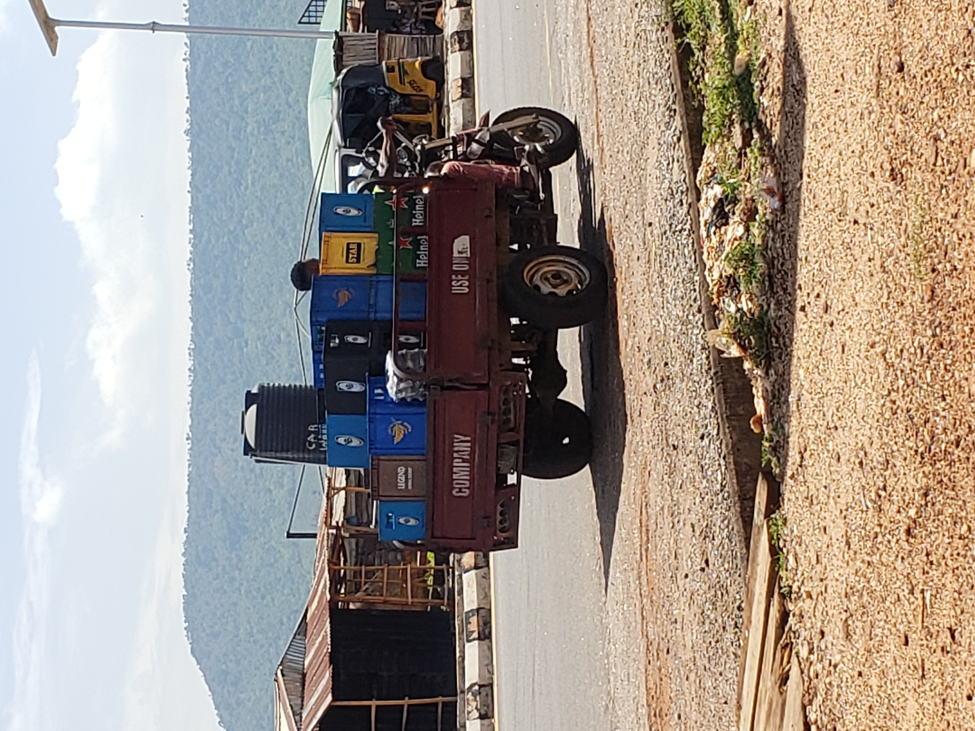 A maroon-colored, three-wheeled motor vehicle is heavily loaded with stacked crates of various beverages, including "Star," "Heineken," and "Legend," and is parked on a paved road in Adankolo, Nigeria. The vehicle, labeled "COMPANY" on its rear, appears to be transporting a significant quantity of bottled drinks. A black water tank is mounted behind the driver's seat. The background shows a hazy mountain range under a partly cloudy sky. A building with a corrugated metal roof borders the road. A narrow strip of vegetation grows along the edge of the pavement. A second yellow motor vehicle can be seen parked further along the road, past the main vehicle.