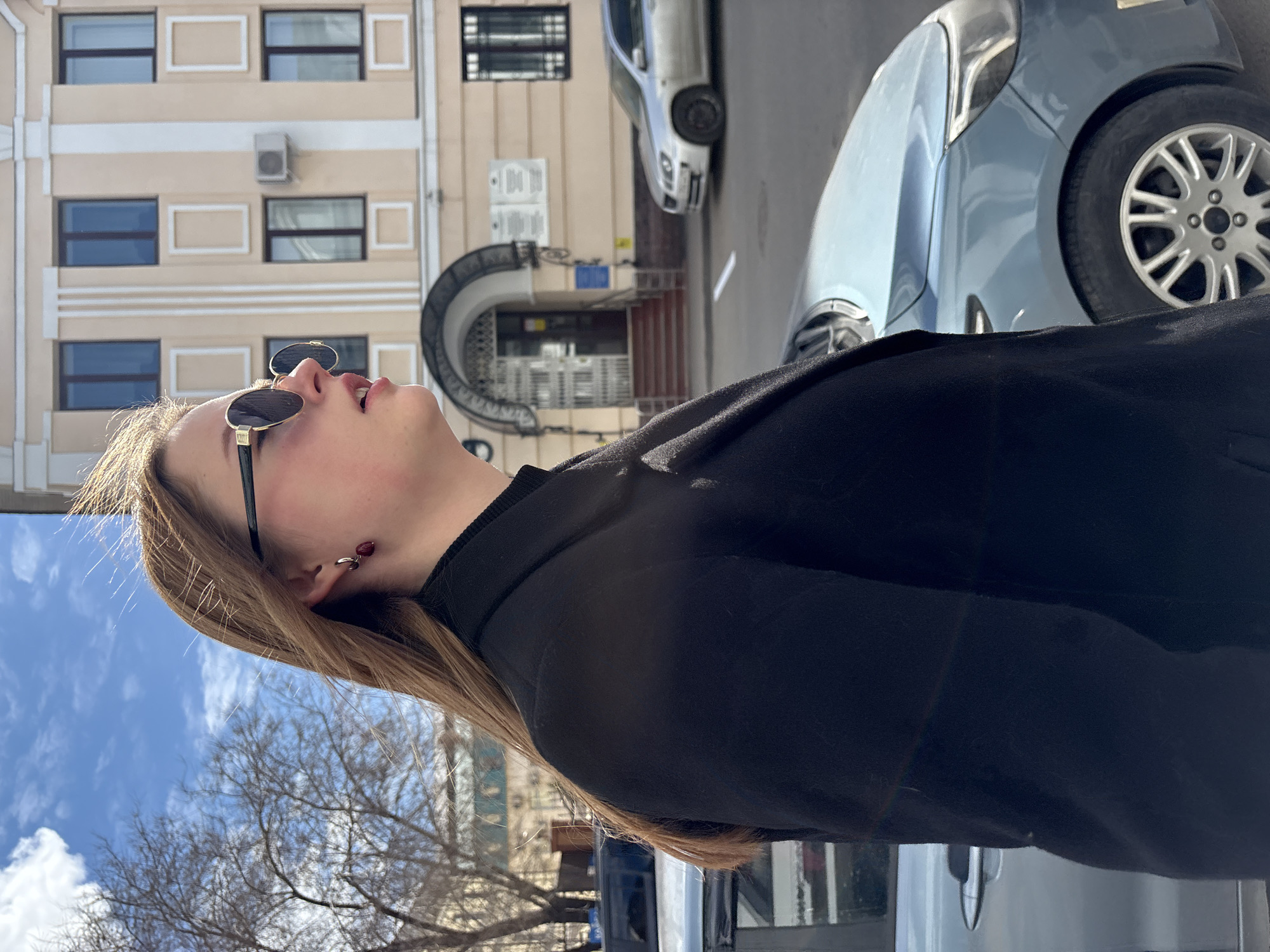 A low-angle shot depicts a woman with blonde hair and sunglasses looking upwards. She is wearing a black coat. In the background are two parked cars and a multi-story building with windows and arched entryways. Bare trees are visible. The location is Kazan, Russia, based on architectural style.
