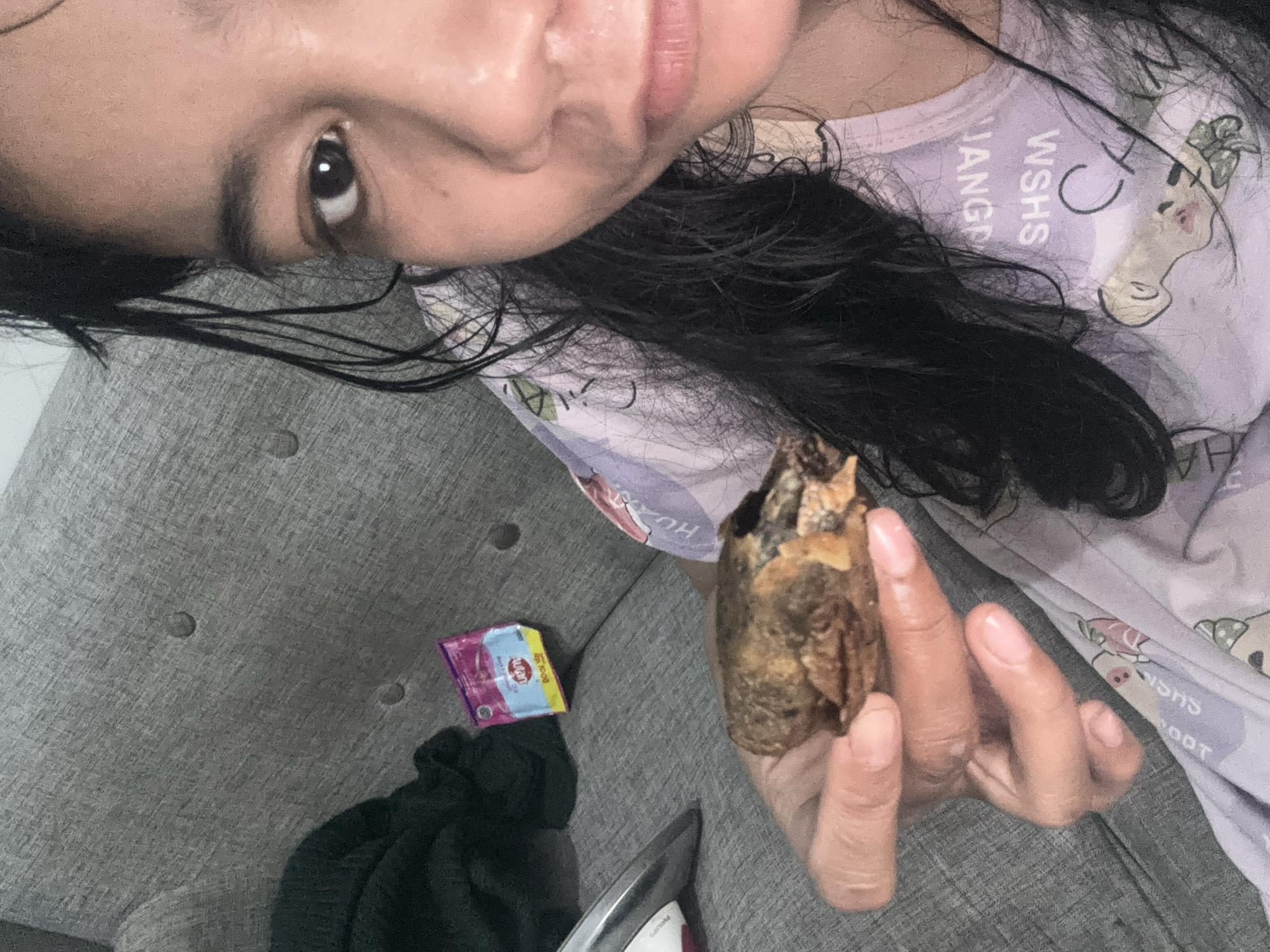 A woman is holding a piece of fried food in her hand. She is wearing a purple shirt with cartoon characters and text. The background is a gray couch, and there is a packet of snacks on the couch. The image appears to be a selfie, taken indoors, likely in a home setting. No specific location within North Jakarta, Indonesia, can be determined from the image alone.