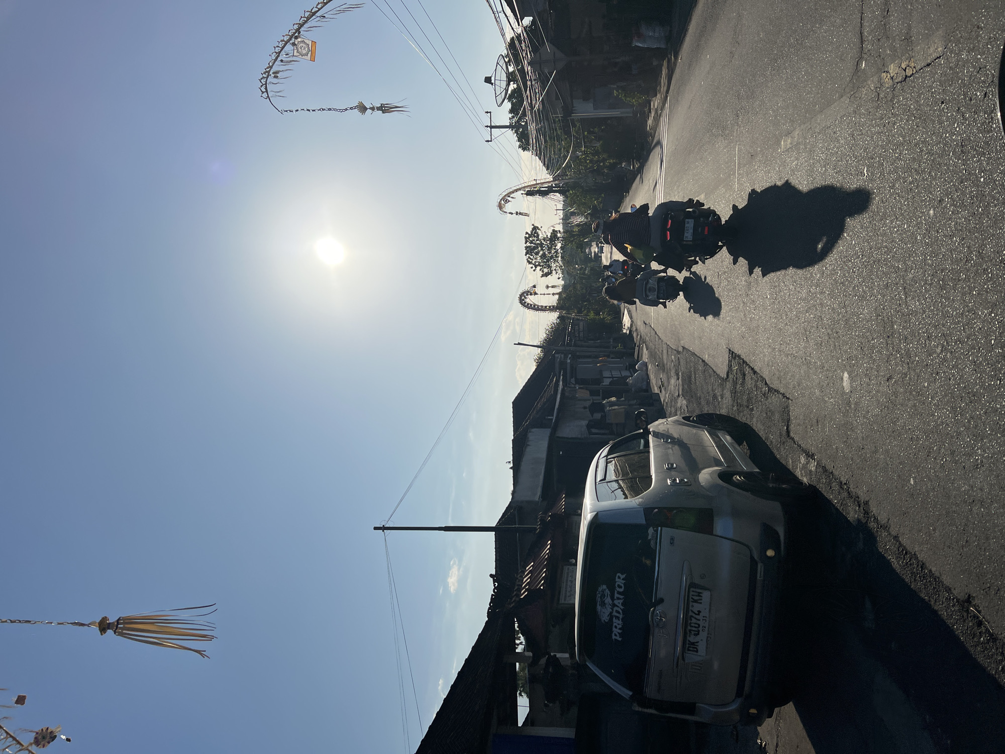 An outdoor, eye-level, full shot captures a street scene in Kubu, Indonesia, on a clear day with the sun high in the sky. The scene is dominated by a road stretching into the distance, flanked by buildings and overhead power lines. A silver sedan is parked on the left side of the road, its rear facing the camera, with a "PREDATOR" sticker on the back window and a license plate that reads "DK 4074 KW". Several people are riding motorcycles down the road, their forms silhouetted against the bright sun. The motorcycles are heading away from the camera. On either side of the road, arched structures with hanging decorations are visible, suggesting a festive or religious occasion. The sky is a brilliant blue with a few wispy clouds. The overall impression is one of a typical day in a bustling Indonesian town, bathed in the warm light of the sun.