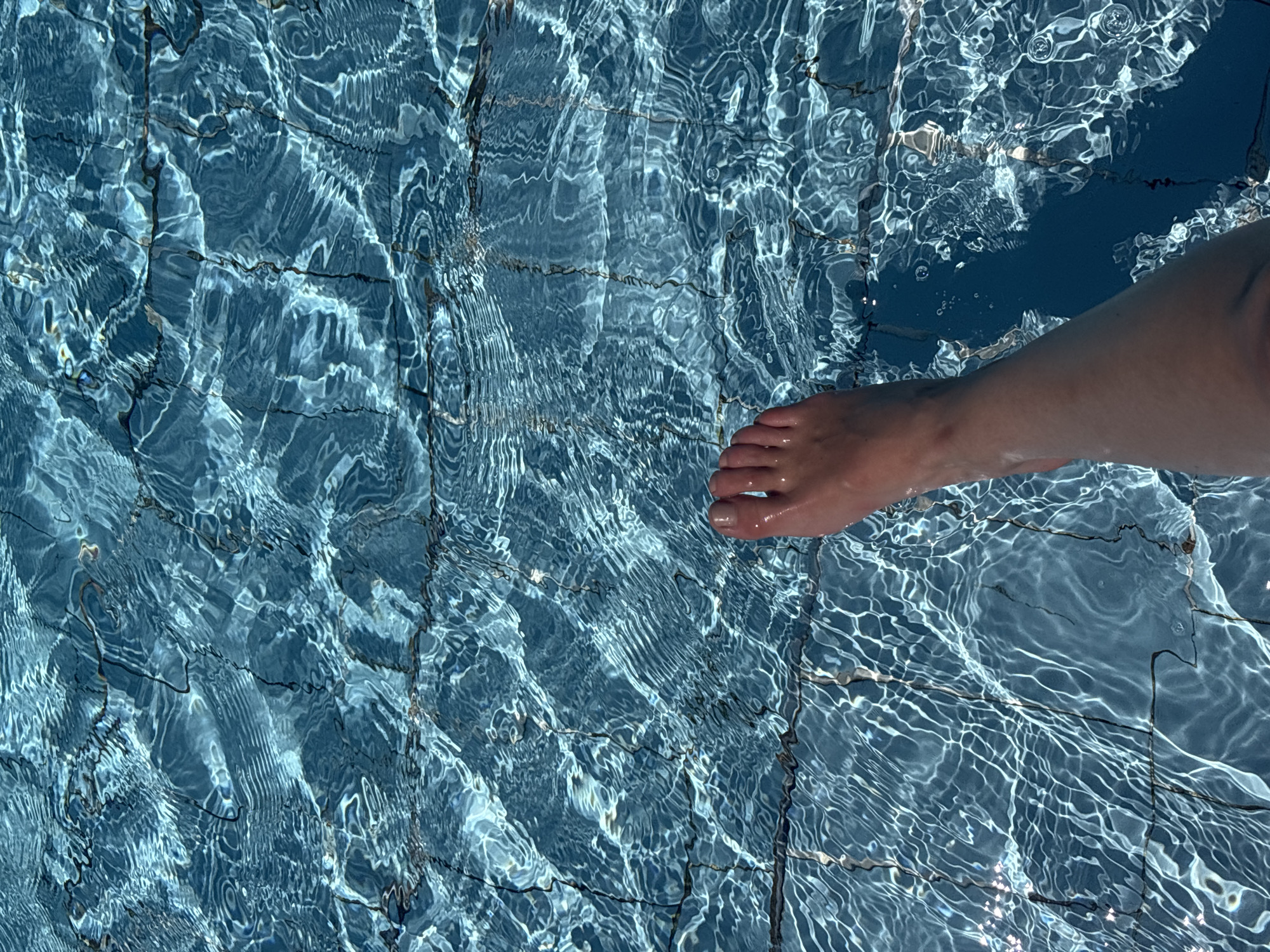 The image depicts a partially submerged foot in a tiled pool. The water is clear with refracted sunlight creating a shimmering effect. The foot, pale in tone, is positioned on the right side of the frame. The pool tiles are light blue, separated by dark grout lines. The location is Unknown, Unknown.