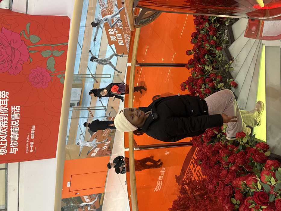 The scene depicts the interior of a multi-level commercial building, likely a shopping mall, located in Jiangjin, China. A dark-skinned female, appearing to be in her 20s or 30s, is positioned on a curved staircase in the foreground. She wears a light-colored beanie, a black puffer jacket, light grey sweatpants, and light-colored sneakers. Her left foot is flat on a step, and her right leg is bent with the foot raised, creating a posed stance. She smiles directly at the camera. The staircase and adjacent areas are densely decorated with artificial red roses. Transparent orange panels serve as railings or architectural elements for the staircase.
In the background, on a lower level of the building, several individuals are visible. One male, dressed in dark clothing, stands near a small child. Further back, two adults are observed near an entrance or passageway, one holding a red object. The lower level features light-colored flooring and orange walls. Large graphic cutouts or murals on these walls depict figures in various poses, including two individuals fencing and one in a dynamic, possibly dancing, posture. Below the fencing figures, the English phrase "BRAVE SELF-CONFIDENCE" is displayed. Above the lower level, a large red banner stretches across the ceiling, adorned with illustrations of pink roses and Chinese characters. Below these characters, the English phrase "EQUALIFY FOR ALL" is printed. The area is well-lit by interior lighting.
