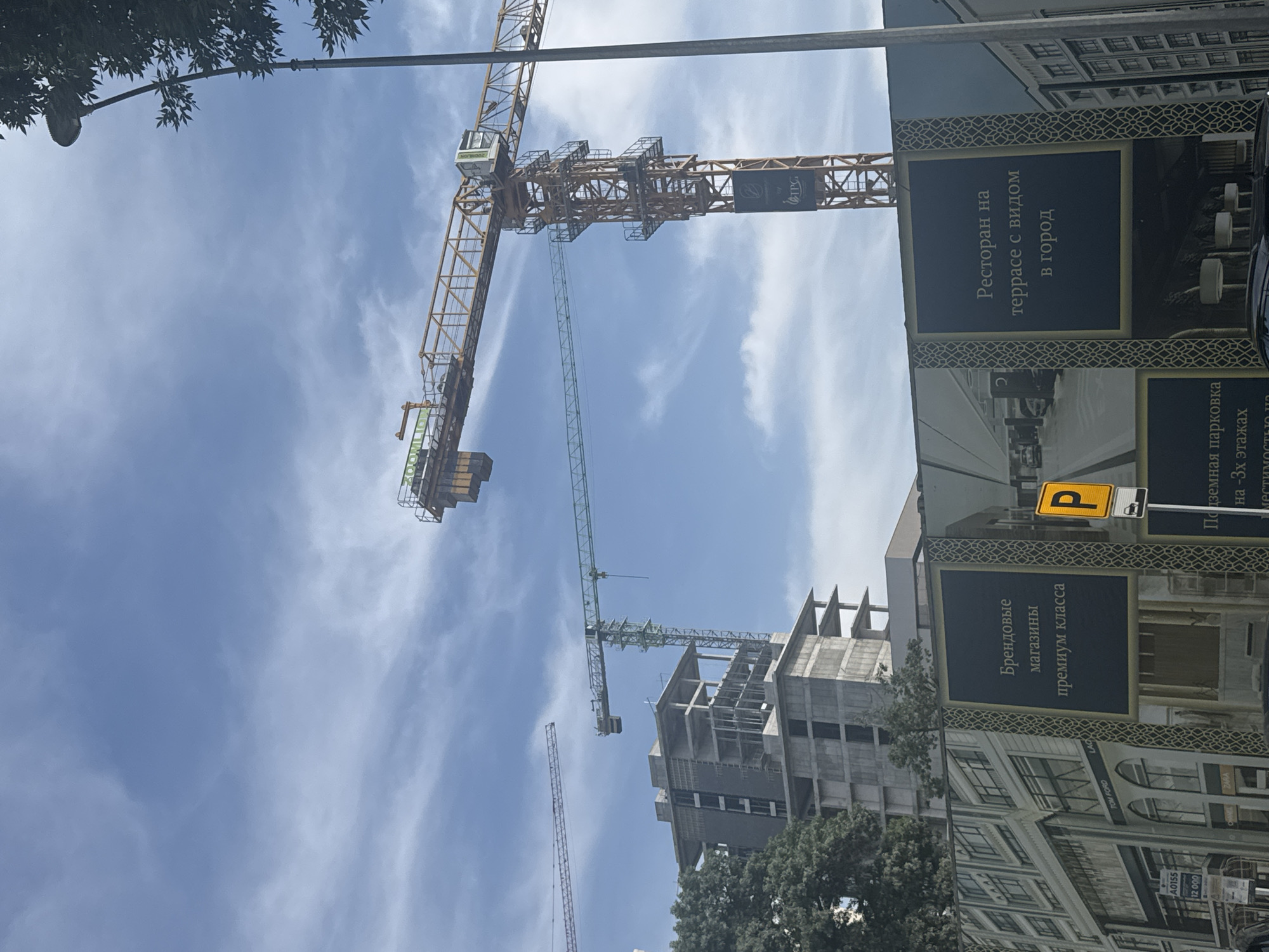 The image shows a construction site in Tashkent, Uzbekistan. Several tower cranes are visible in the frame, suggesting ongoing building activity. There is an advertisement for a restaurant with a view in the city and premium brand stores. A parking sign is also visible. The buildings are concrete and have exposed rebar. The sky is blue with scattered clouds. There are trees in the lower part of the frame. The signs are in Russian.