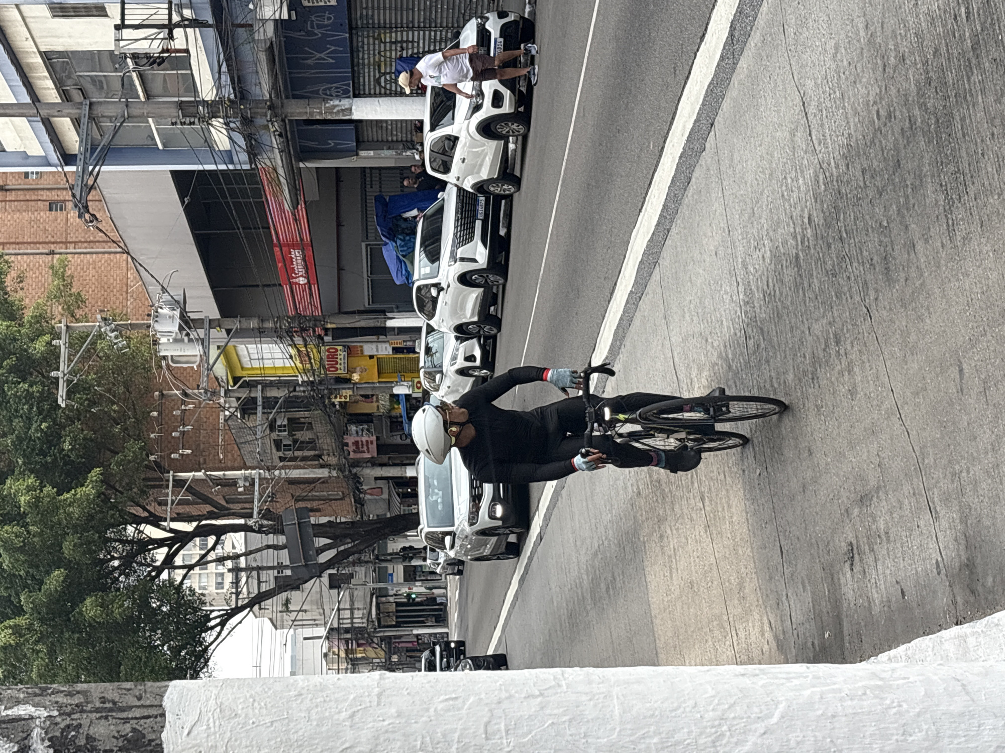 The image depicts a street scene in São Paulo, Brazil. In the foreground, a cyclist wearing a black full-body suit, white helmet, and gloves rides a bicycle on a paved sidewalk. The cyclist is looking to the right. Behind the cyclist, the street is lined with parked white cars, including a white Volkswagen Nivus and a white Fiat Argo. Further down the street, there are buildings with shops and signs, including a Santander bank branch. Power lines and trees are visible in the background, characteristic of a developed urban environment. The overall lighting suggests it is daytime.