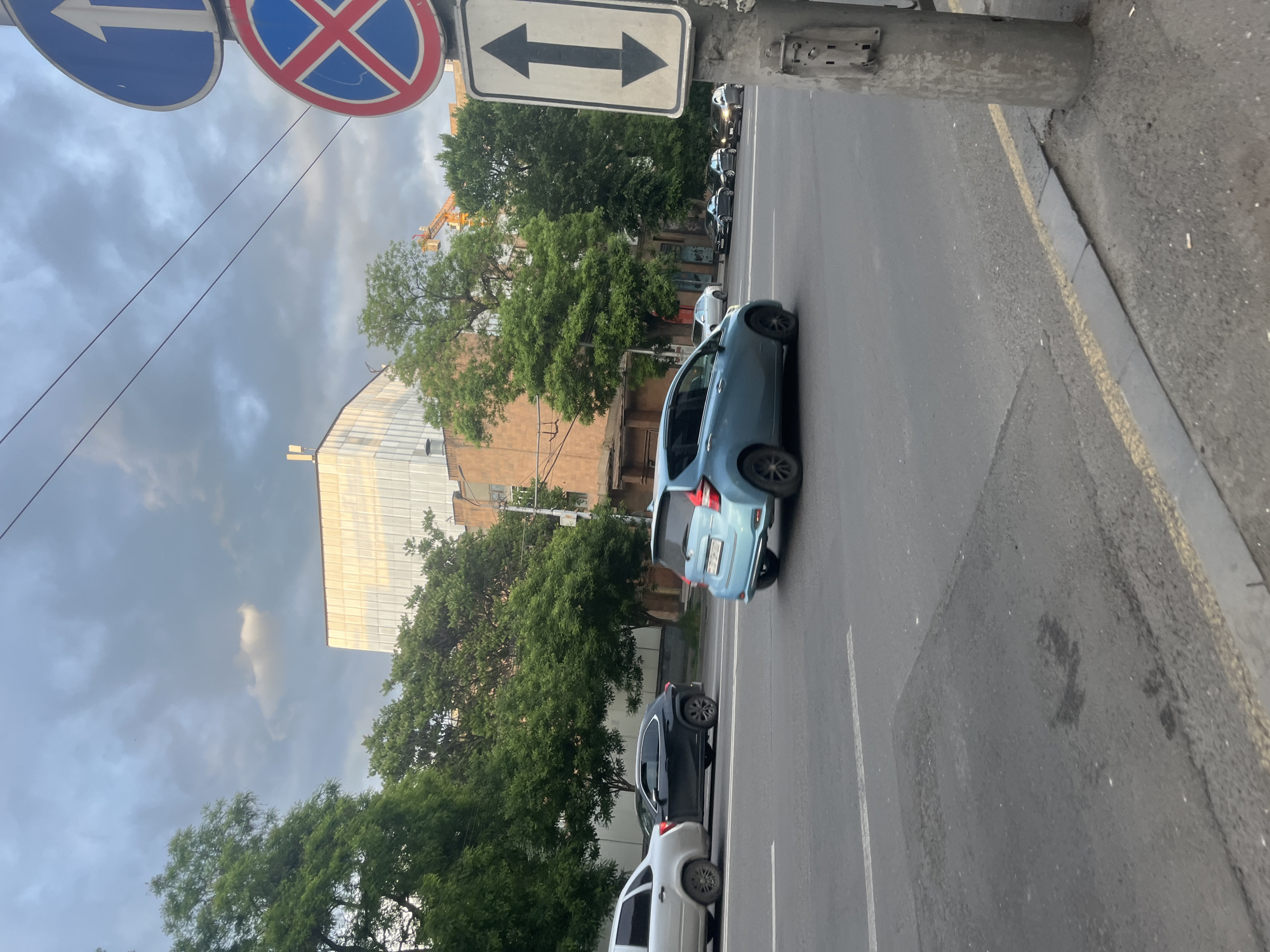 The image depicts a street scene in Yerevan, Armenia, identifiable by architectural details and license plates. A light blue hatchback car is in the foreground lane moving right, followed by two parked cars in the same lane, dark and white. The street is divided by a solid double white line. A sidewalk lines the right edge of the frame. Above are the signs for direction and a stop. Trees and an older building are visible in the background. The sky is overcast. A crane is visible in the background.