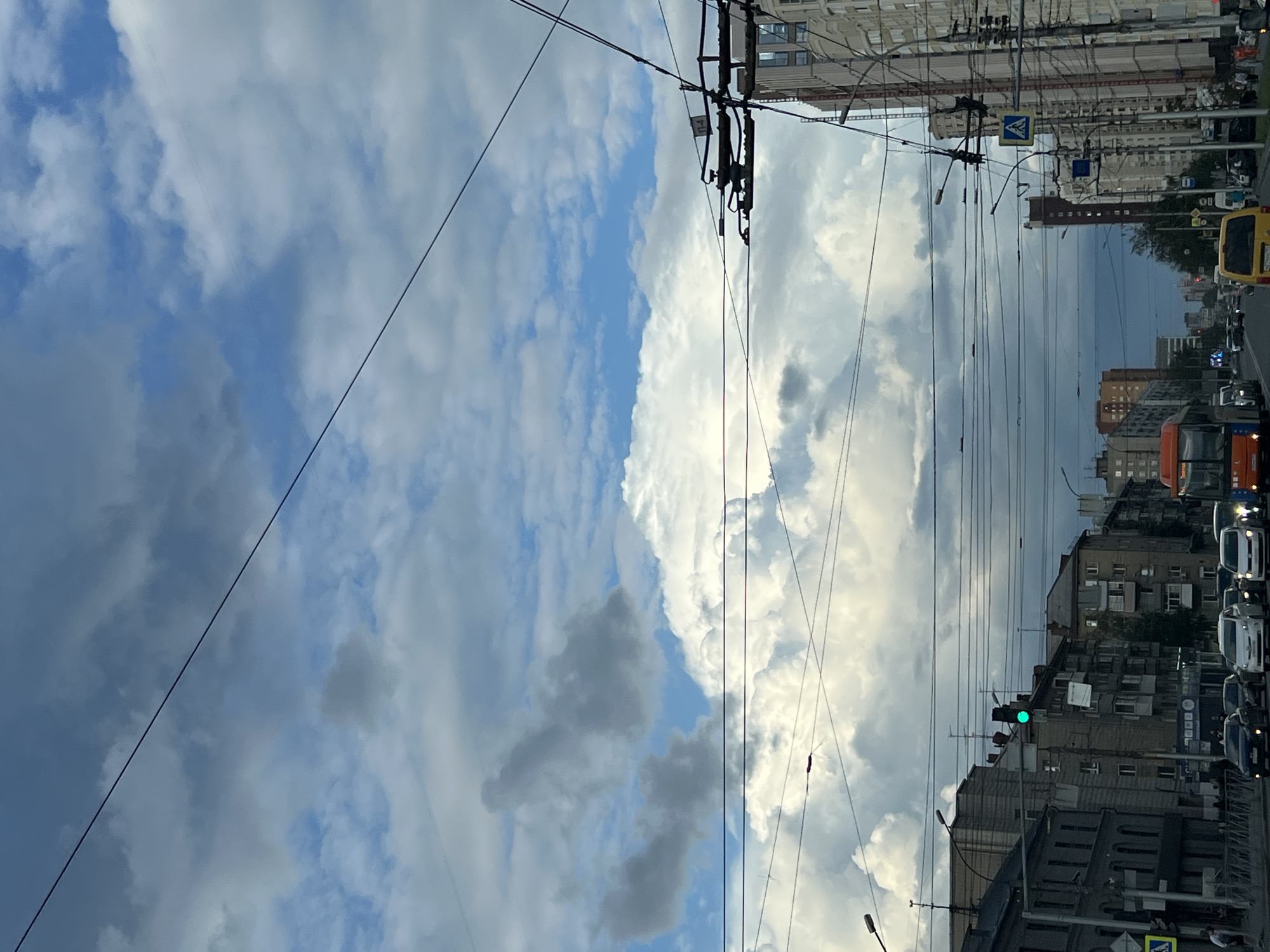 The image depicts a street scene in Novosibirsk, Russia, under a dramatic, cloudy sky. The perspective is from a vehicle traveling down a wide, multi-lane road lined with apartment buildings on both sides. Overhead, a complex network of trolleybus wires crisscrosses the sky, punctuated by traffic lights and street lamps. Several cars and a yellow van are visible in traffic, along with an orange bus in the distance. A green traffic light is illuminated. The architecture of the buildings suggests an urban environment. The overall impression is of a typical day in a Russian city, with the emphasis on the dynamic weather patterns overhead.