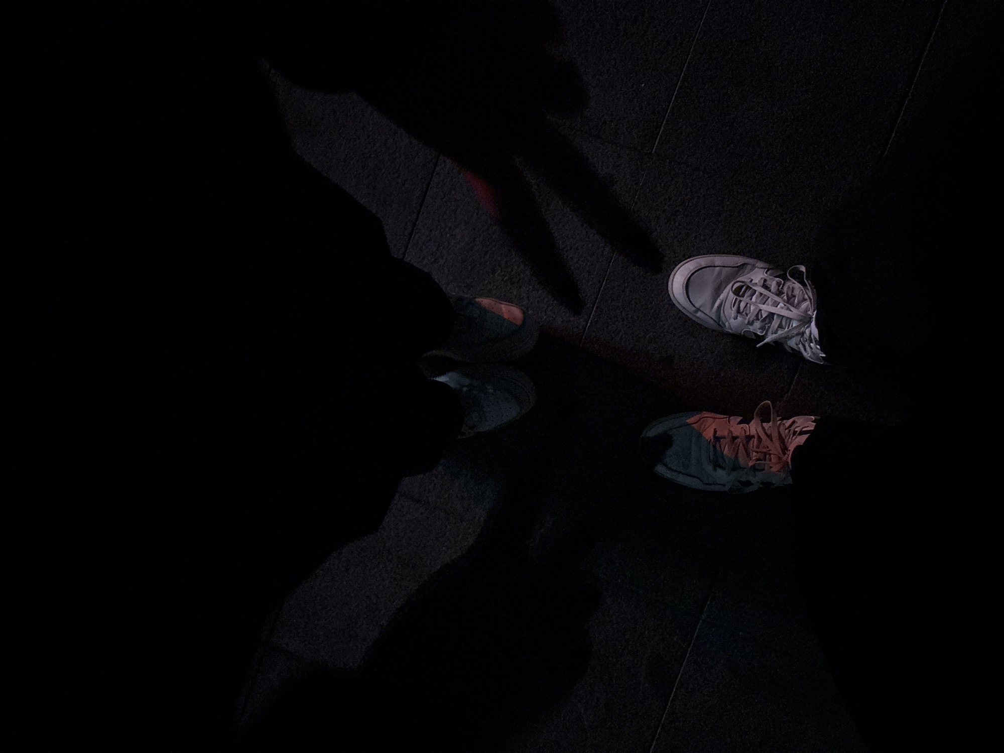 The image shows a dimly lit scene, likely indoors on a dark tile floor. There are three pairs of athletic shoes visible. The first pair, located near the top right, is a light color. Another pair can be seen in the bottom right corner. A third pair is located in the upper-left portion of the image, and partially obscured by shadow. The shadows of three people surround the shoes, one making the peace sign. The overall darkness suggests a low-light environment. There is no information available to indicate this image was taken in Burgas, Bulgaria.
