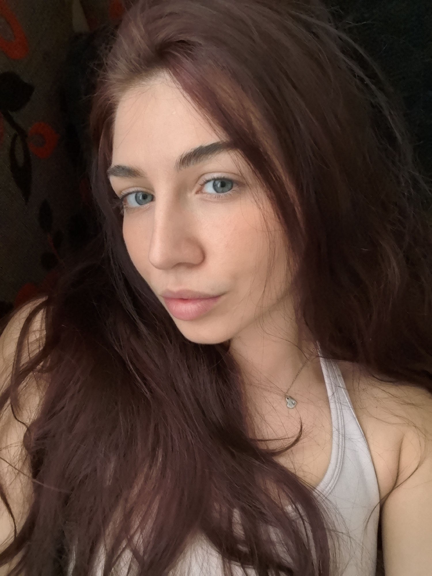A medium shot depicts a Caucasian woman with long, auburn hair, facing slightly left of the viewer. Her eyes are light blue, and her skin has a fair complexion. She wears a simple, light-colored tank top. A delicate silver necklace with a small charm rests on her chest. The background is a blurred mix of dark fabric, suggesting a dimly lit room. No specific location in Stara Zagora, Bulgaria, can be determined from the image.