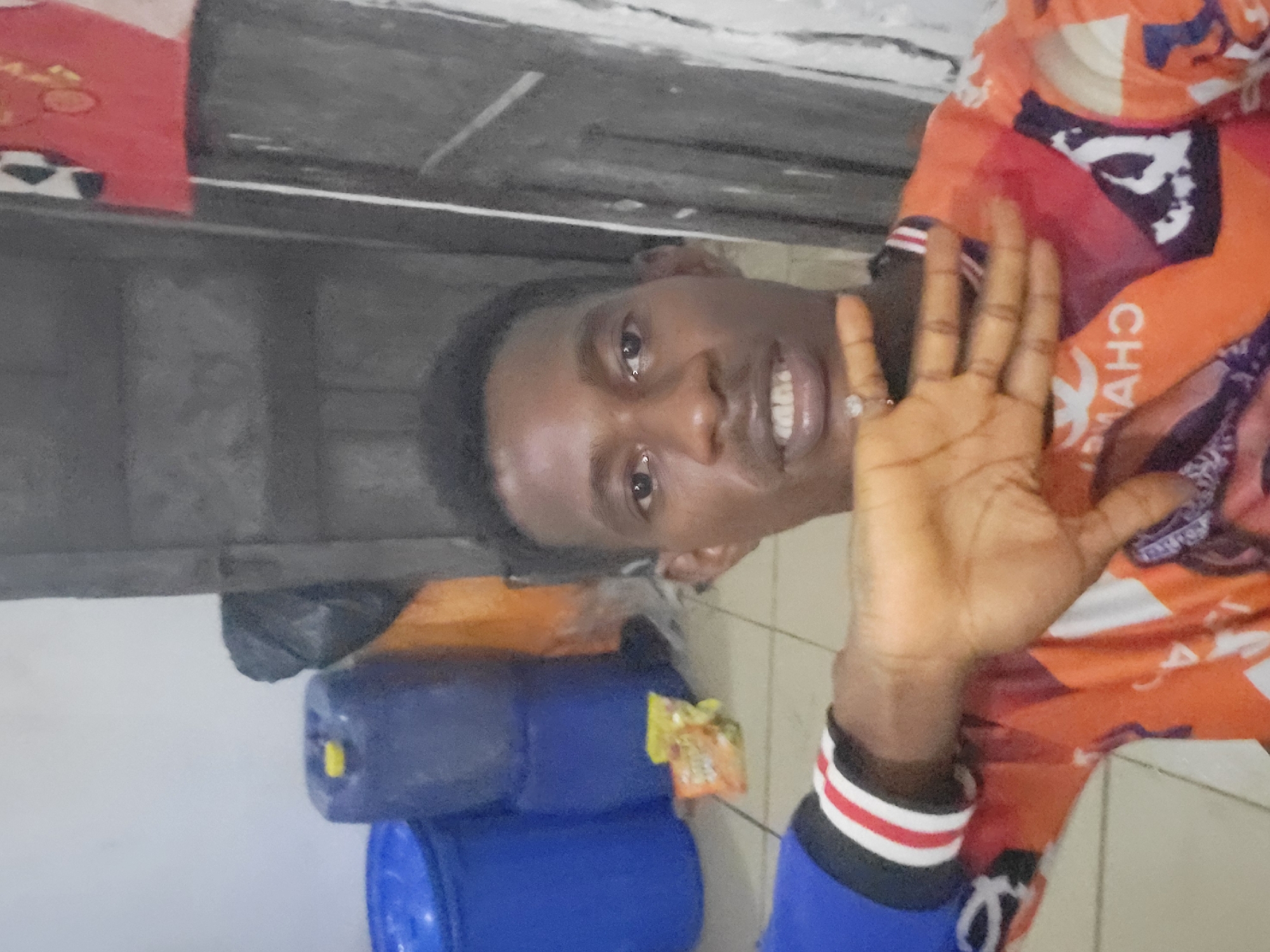 In an indoor setting, a young Black man with short, dark hair and brown eyes takes a selfie. He is wearing a blue long-sleeved shirt with a blue, red, and white striped cuff and an orange t-shirt with a graphic design. His left hand is raised in front of his face, and he is wearing a ring on his left ring finger. Behind him are several large blue barrels, a black trash bag, and an orange object. The wall behind him is a light color, and there is a gray door or cabinet to his right. The floor appears to be tiled.