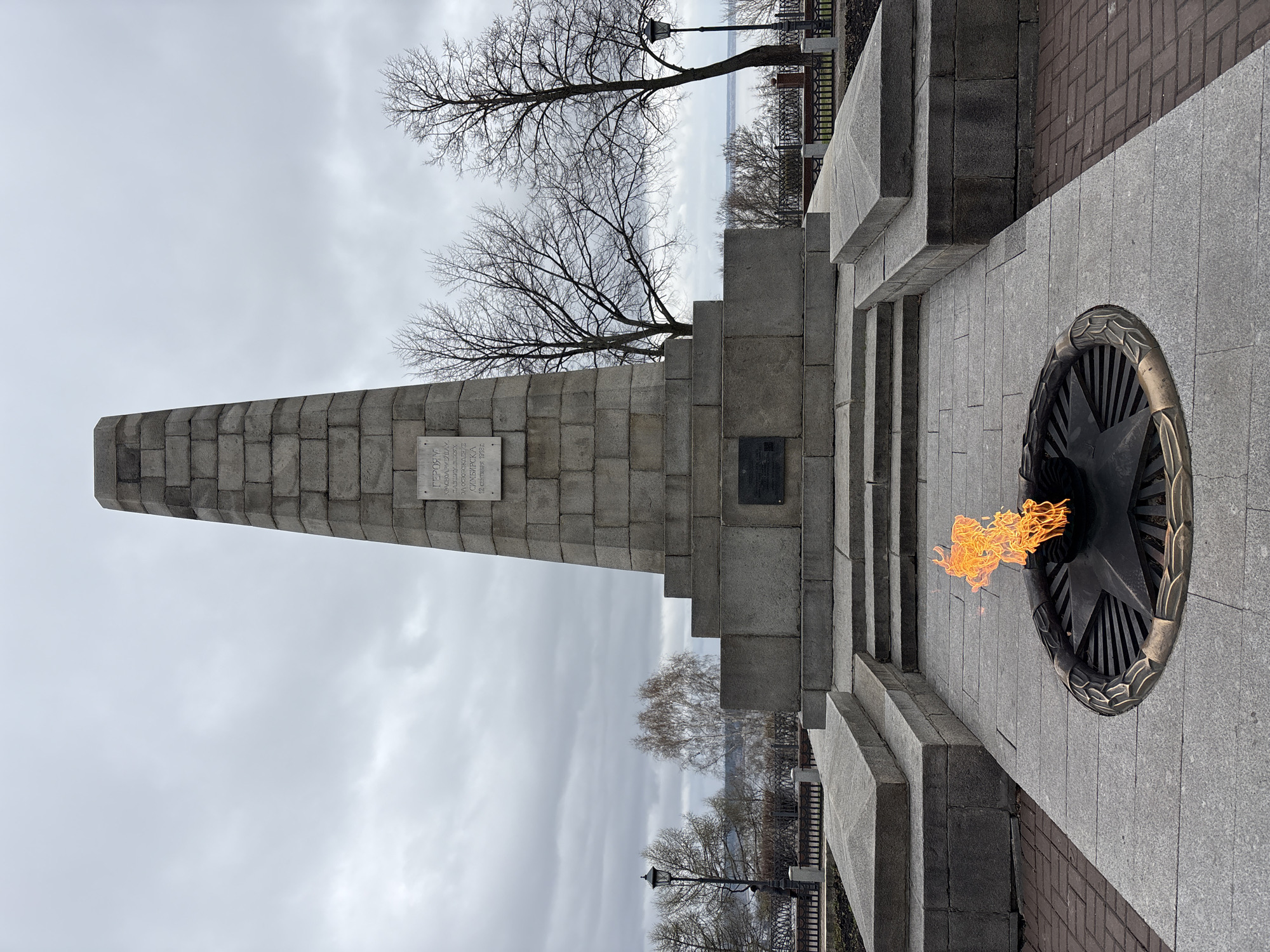The image captures a solemn war memorial on an overcast day in Ulyanovsk, Russia. In the foreground, an eternal flame burns in a dark, star-shaped metal base embedded in light gray paving stones. To the left, a substantial stone monument rises, composed of a stepped base leading to a tall, tapering obelisk constructed from large, rectangular stone blocks.
On the obelisk, a light-colored plaque displays Cyrillic text. The inscription reads: "TO HEROES WHO SACRIFICED THEIR LIVES FOR FREEDOM." It further specifies "SIMBIRSK, 12 SEPTEMBER 1918" and "OCTOBER 1918," referencing significant historical events from the Russian Civil War in Simbirsk (the former name of Ulyanovsk). A second, darker plaque with indistinct text is visible on a lower section of the monument's base.
The background features bare trees, suggesting a late autumn, winter, or early spring season. Beyond the trees, a wide body of water, likely a river, is visible under a uniformly cloudy, gray sky. A traditional black lamppost and a decorative metal railing are present along a pathway near the water's edge. The overall scene evokes remembrance and historical significance, with no people visible.