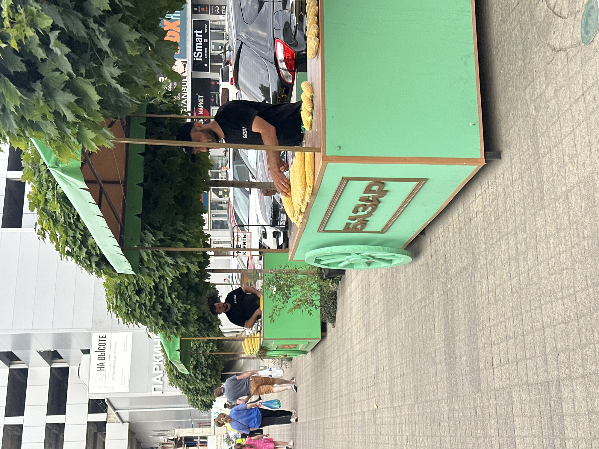 In Stavropol, Russia, on a paved sidewalk lined with trees, two mobile corn stands are set up. The foreground stand is bright green with a wooden trim, displaying piles of corn on the cob. A man in a black t-shirt and a black cap, with "BAZAR" printed on his shirt, stands behind the counter, arranging the corn. Behind him, a second green corn stand is visible, also with corn displayed, and a vendor in a black t-shirt stands behind it. In the background, pedestrians walk along the sidewalk, and a multi-story building with the sign "НА ВЫСОТЕ" (which translates to "At Height") and "ПАРКИНГ" (Parking) is visible. Several cars are parked in front of shops with signs including "Istanbul," "iSmart," and "MAPKET." The weather appears overcast.