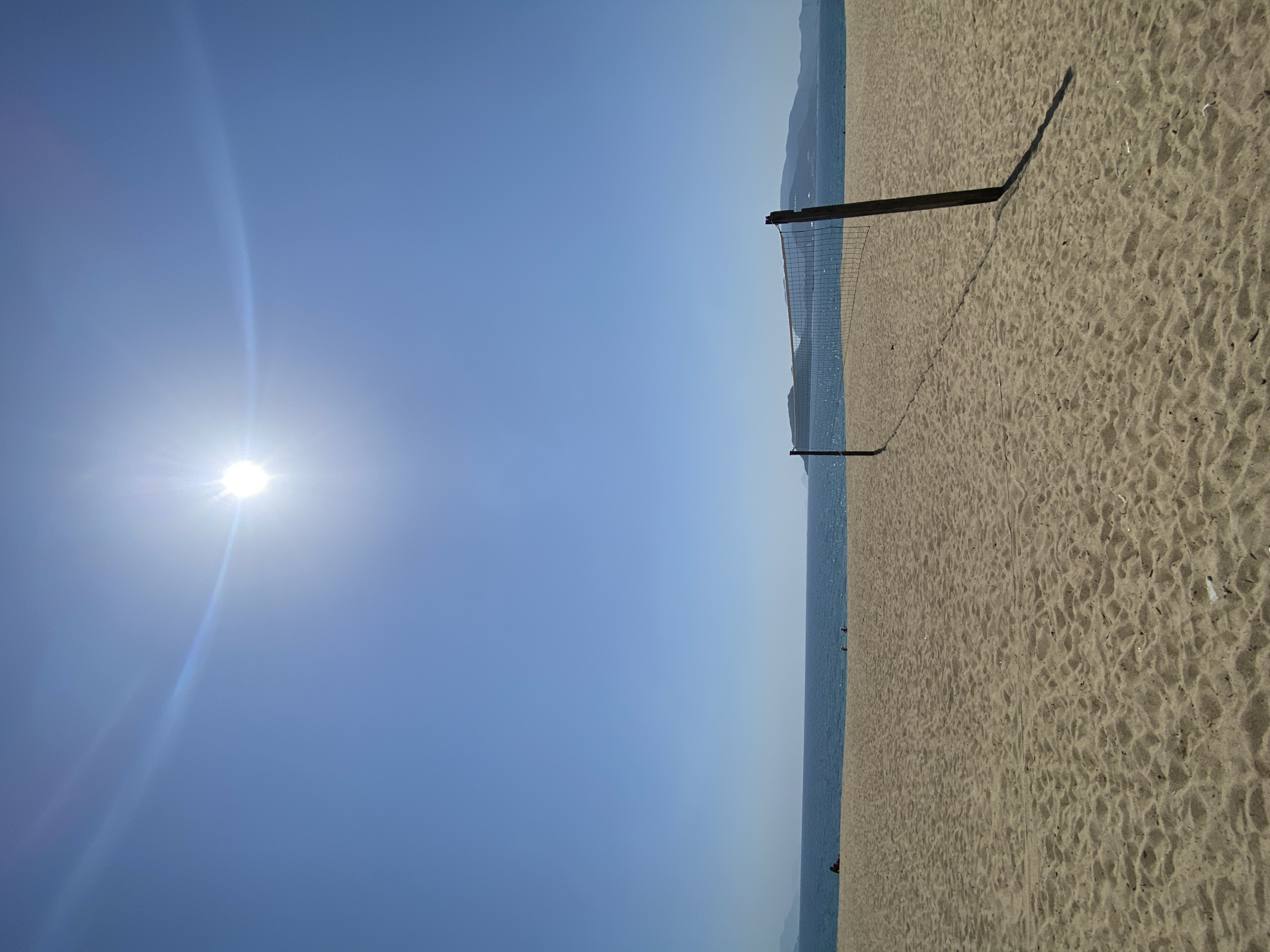 The image depicts a serene beach scene in Nha Trang, Vietnam, under a clear, bright blue sky. The sun, positioned high in the sky, casts a strong glare and a lens flare effect, indicating midday or early afternoon. The foreground is dominated by a vast expanse of light brown sand, marked with subtle ripples and indentations. In the middle ground, a volleyball net stands tall, with two wooden posts supporting its mesh structure. The net stretches across the sandy expanse, casting a distinct shadow on the sand due to the harsh sunlight.
Beyond the volleyball net, the calm, blue ocean stretches to the horizon, meeting the sky in a soft, indistinguishable line. In the distance, along the horizon, several hilly islands or peninsulas are faintly visible, their shapes softened by atmospheric haze. A few scattered figures can be seen on the beach and near the water's edge, suggesting the beach is not entirely deserted, but there are no immediate interactions or activities captured. The overall atmosphere is one of tranquility and leisure, typical of a sunny day at the beach in Nha Trang.