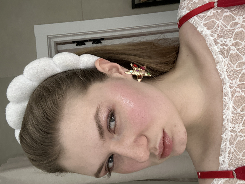 A fair-skinned person wearing a white, lace top with red straps is angled in a portrait. The person has light brown hair partially covered by a white, cloud-like headband. They wear a star-shaped earring adorned with multiple colored jewels. Their skin has a subtle blush, and their lips are glossy. The background shows a neutral-colored wall and a portion of a white door frame.