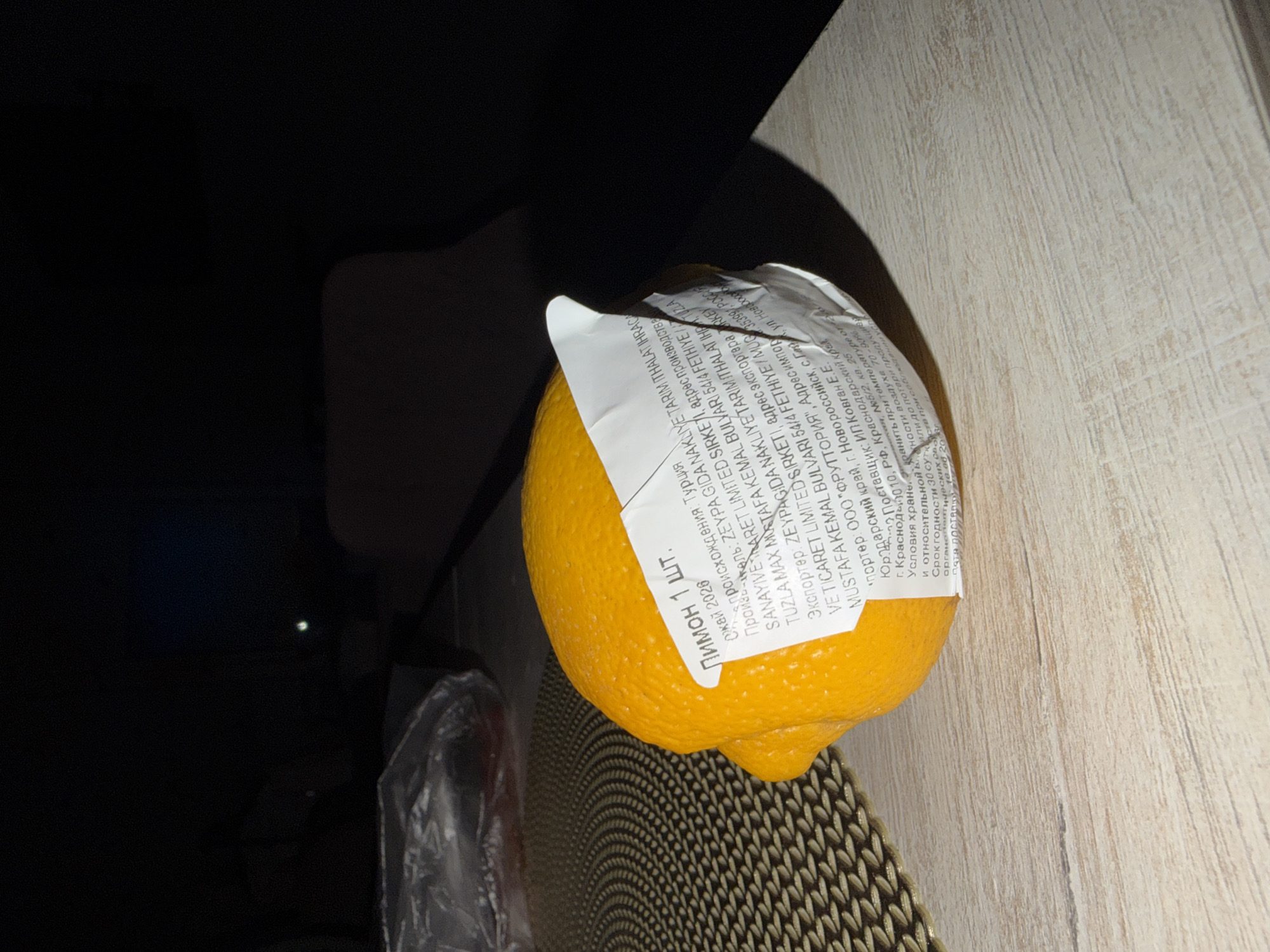 A whole, bright orange lemon is positioned on a textured, light-colored table. A white label is attached to the lemon, displaying text in Russian and English, including "Лимон 1 ШТ." (Lemon 1 PC.) and origin information about "ZEVPAGIDA NAKLYVE TARIM THALAT IHRA". The background is dark and out of focus, with a hint of a light source in the upper right quadrant. The image appears to be taken indoors, with artificial lighting casting shadows. There are no people or animals visible in the frame. The city of Sirius, Russia, is not depicted in this image; the image is focused solely on the lemon and its label.
