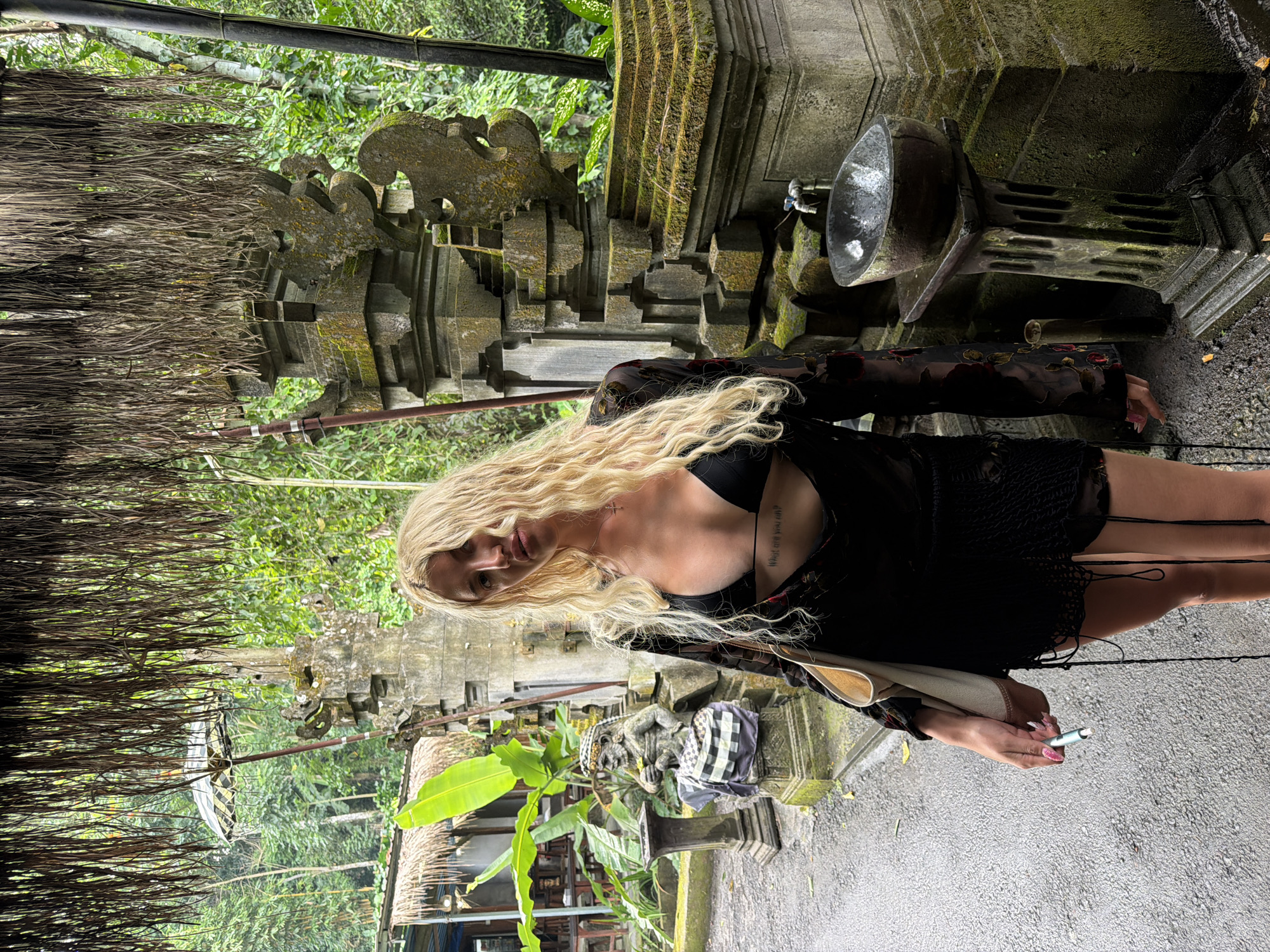 A young woman with long, wavy blonde hair stands in front of a stone structure covered in moss, holding a cigarette in her right hand and a bag in her left. She is wearing a black bikini top with a sheer, long-sleeved floral cover-up and a black skirt. The background features lush greenery, a thatched roof, and a stone statue. The scene appears to be in a tropical outdoor setting, possibly a temple or garden in Gianyar, Indonesia, given the Balinese architectural style of the stone carvings and the statue.