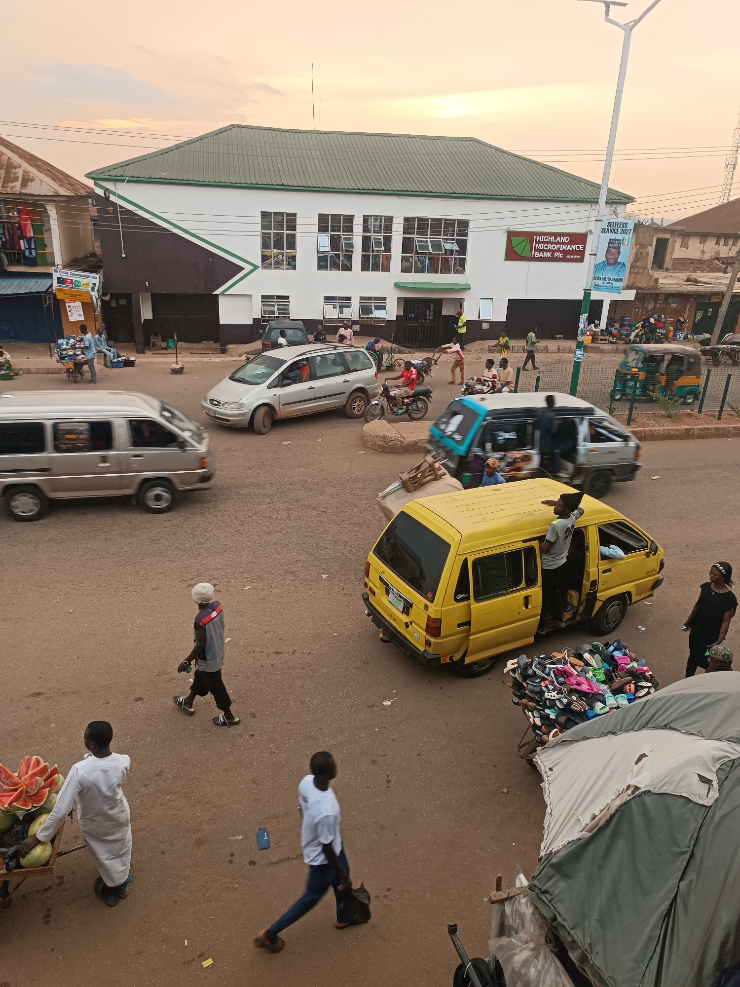 The image depicts a busy street scene in Bukuru, Nigeria. The scene is shot from an elevated perspective, possibly from a building overlooking the street. Multiple vehicles are present including vans, a car, a motorcycle, and an auto rickshaw. People are walking, standing, and interacting near the vehicles. A yellow van is prominent, with a person partially inside. A vendor is selling watermelons from a cart. A Highland Microfinance Bank Plcl building is visible.