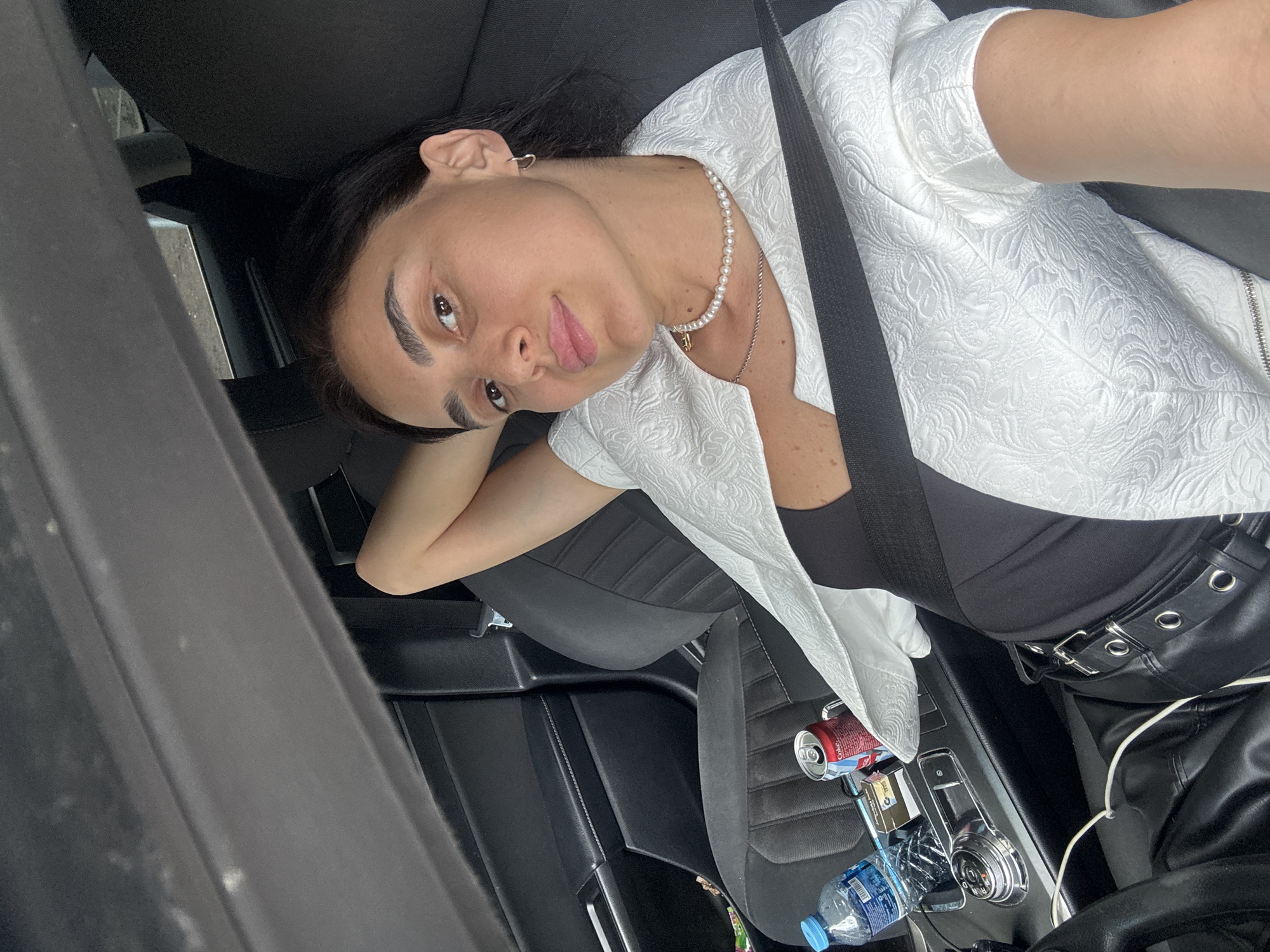 A woman is sitting in the passenger seat of a car in Tbilisi, Georgia, with her arm behind her head. She is wearing a white patterned jacket over a black top and black pants with a belt. A bottle of water and a can of Coca-Cola are visible on the center console. The car's interior, including the seats and dashboard, is dark. The background outside the car is blurred, suggesting movement or distance.