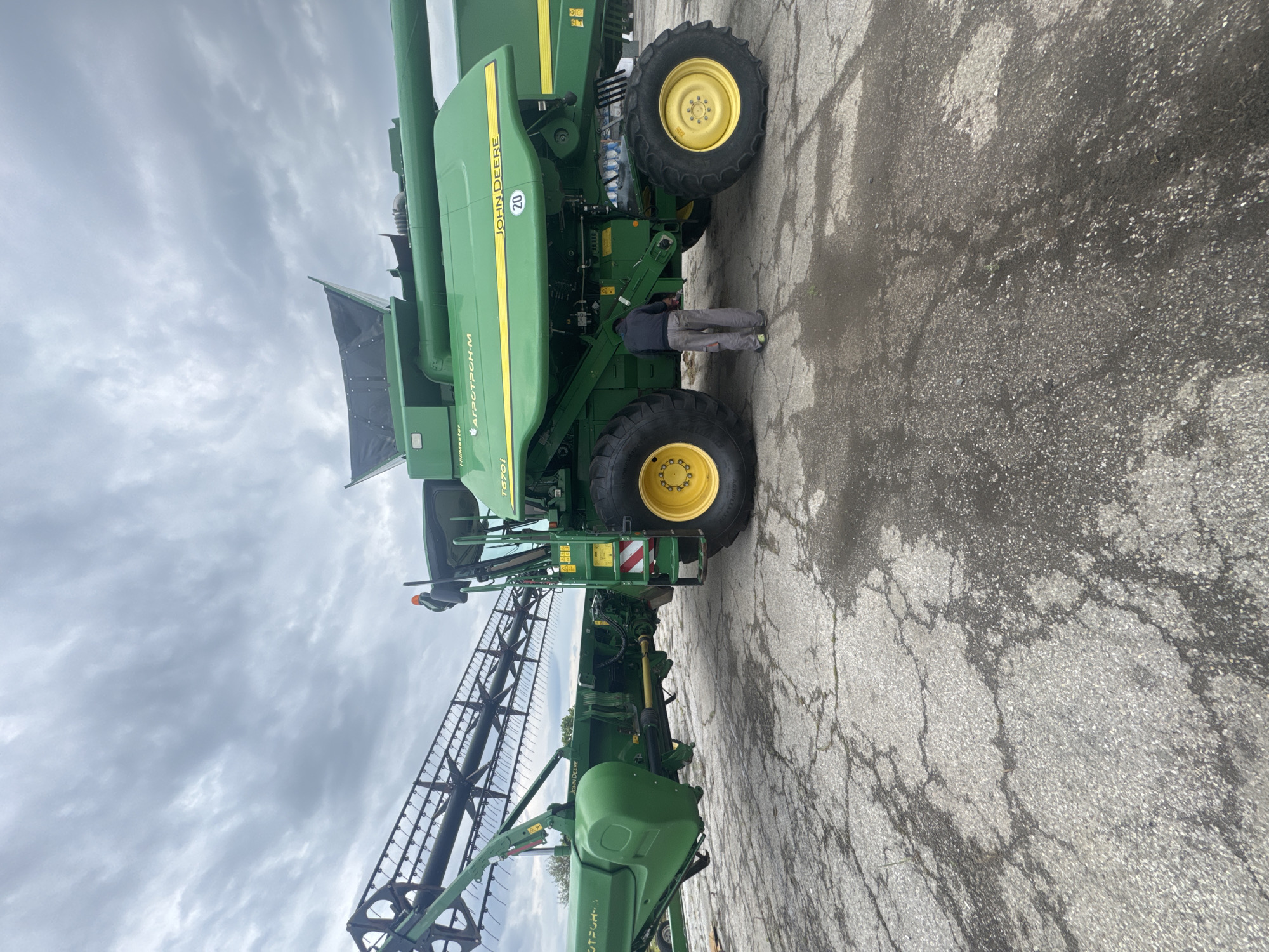 The image shows a green John Deere combine harvester parked on a cracked concrete surface. A person wearing gray pants and a dark jacket is standing near the combine. The sky is overcast. The combine has a header attached. "Agrotron-M" is printed on the side of the combine. The number "20" is visible on the side of the vehicle. Based on the markings on the side of the combine, "АГРОТРОН-М" (AGROTRON-M), the machine is likely in or from Bulgaria, and the image may have been taken in or around Sarnevo.