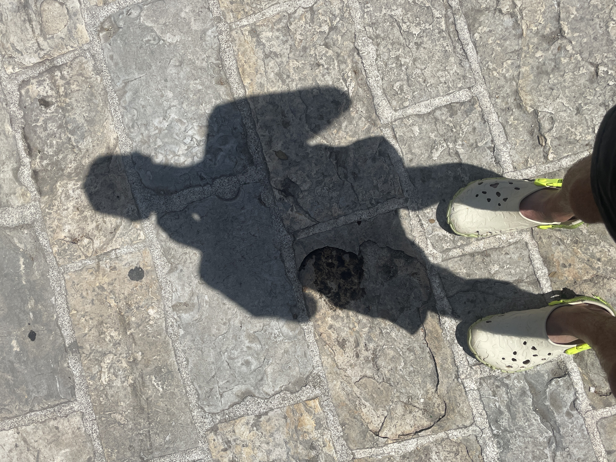 A shadow of a person wearing light-colored clogs with neon green accents is cast on a cobblestone walkway. The shadow displays the silhouette of a person with arms extended outwards. The walkway is made of irregular, weathered stones with visible grout lines. The scene is outdoors, likely during daylight hours, indicated by the strong shadow. The location is described as Unknown, Unknown.