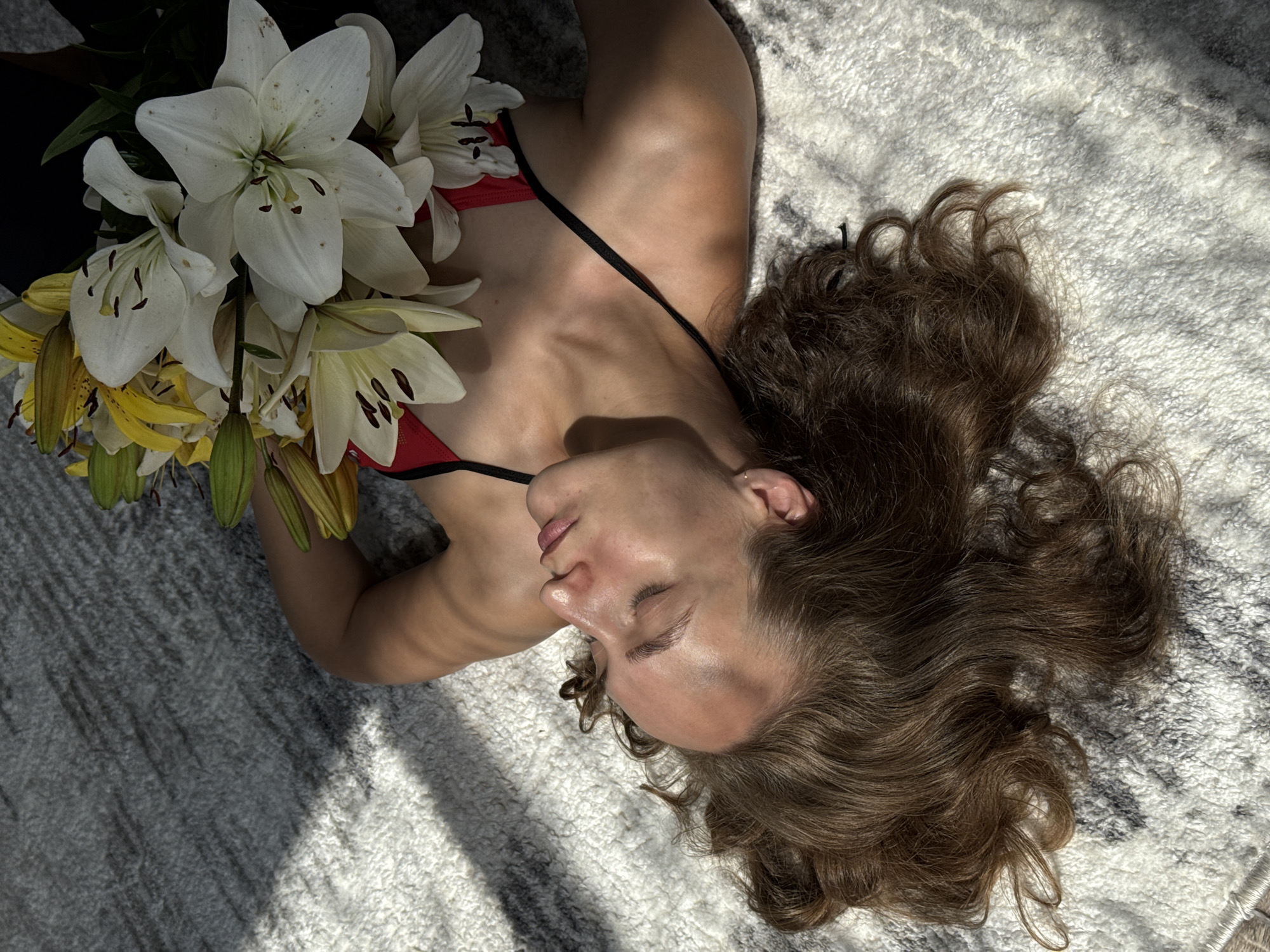 A person with light skin and brown wavy hair lies on their back on a textured rug, on which sunlight casts diagonal shadows. Their head is turned to the left and slightly upwards, with their eyes closed. A bouquet of lilies, with white and yellow petals, rests on their chest. The person is wearing a red top with thin black straps. The background is a rug with a grey and white pattern, and parts of the rug are in shadow. No specific location in Sharkan, Russia is identifiable from the image.