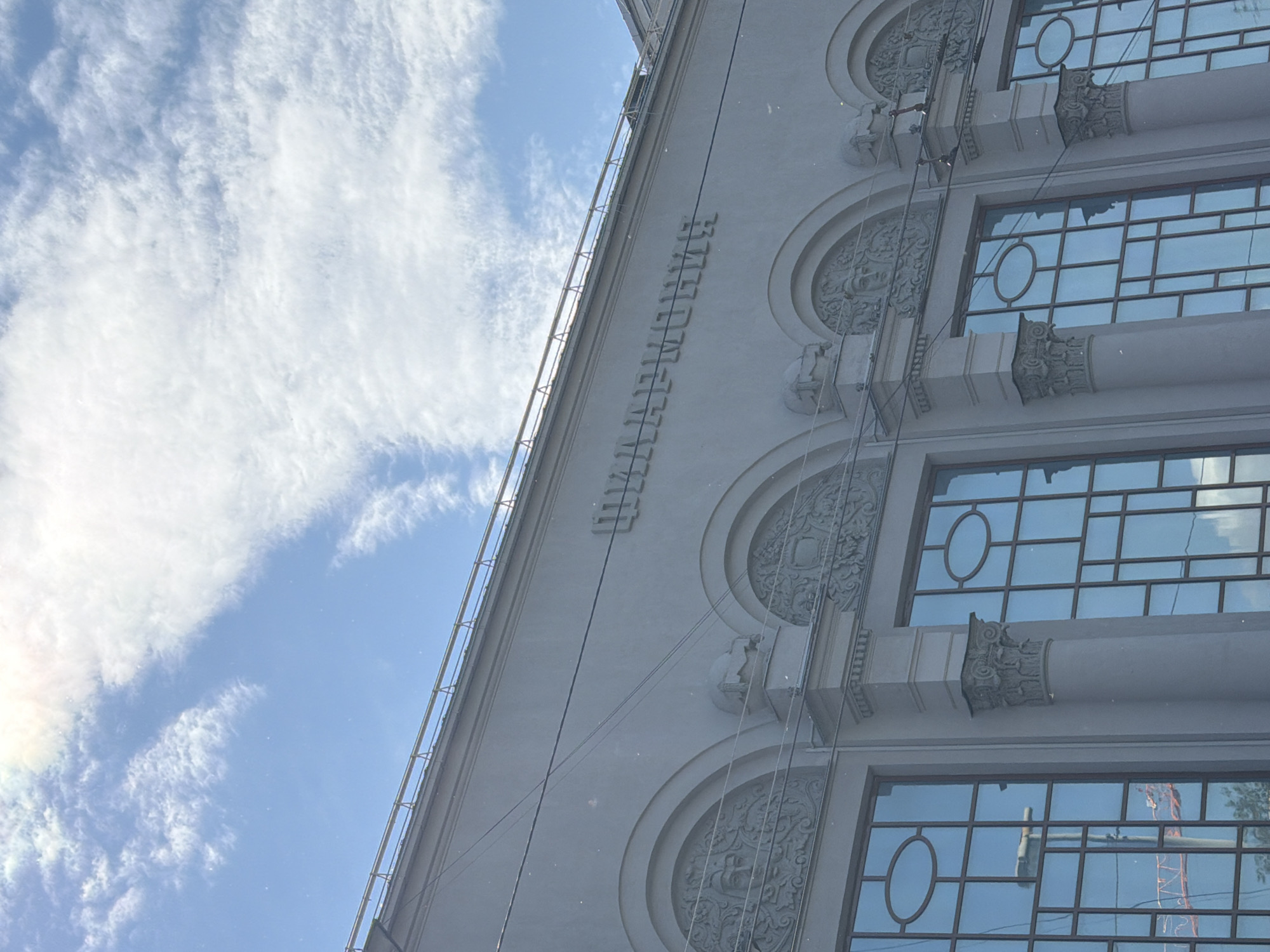 The image shows a close-up, medium-angle shot of the facade of a building with ornate architectural details, located in Yekaterinburg, Russia. The building's facade is a light gray color and features a series of arched windows. Above the windows, there is carved ornamentation, including what appear to be faces within some of the arches. The name of the establishment, "ЦИРК" which translates to "CIRCUS", is embossed in large, stylized letters across the top of the facade. The sky above is a bright blue with scattered white clouds, and sunlight creates a slight lens flare in the upper left corner. Electrical wires are visible stretched across the building. The windows reflect the sky and some surrounding structures, including a red construction crane. The style of the architecture suggests it is a historic building.