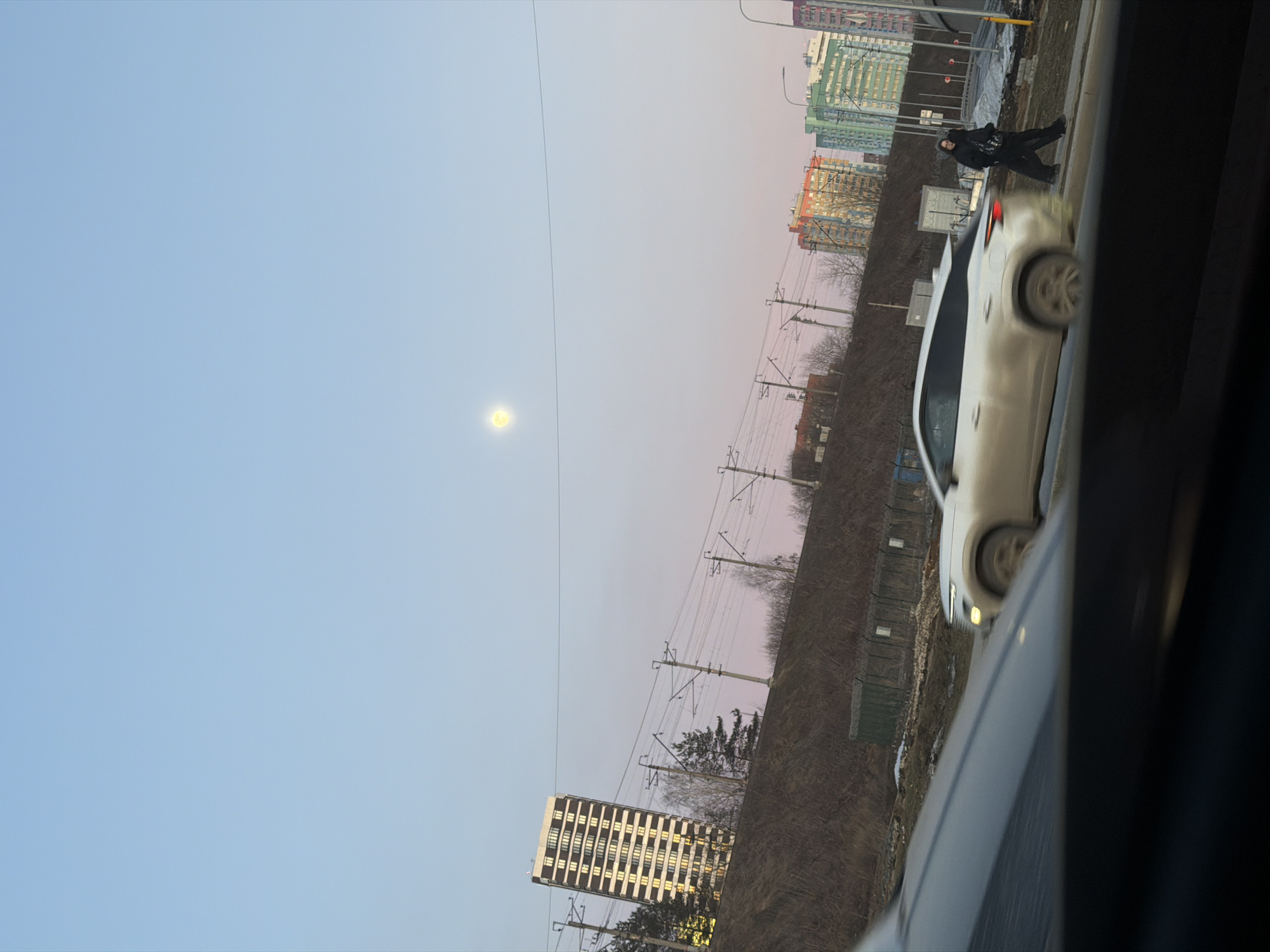 The image depicts a street scene in Perm, Russia, at dusk. A white car is parked on the side of the road. A person in dark clothing is walking on the sidewalk next to the car. In the background, several multi-story buildings and power lines are visible. The sky is a gradient of blue and pink, with a bright orb, possibly the moon or sun, in the upper left. The view is partially obscured by elements of the vehicle from which the photo was taken. A grassy embankment rises up from the road.