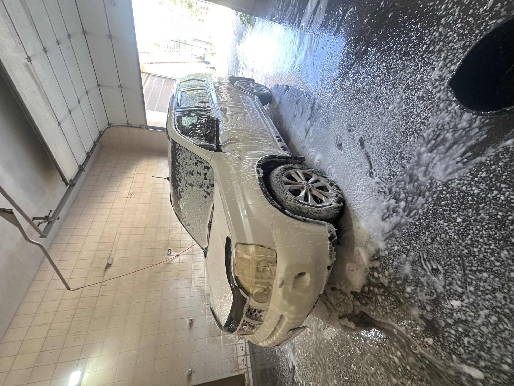The image shows a white SUV covered in soap suds at a car wash. The car is positioned in the center of the frame, angled slightly to the left. The car wash appears to be an open-air or partially enclosed structure with a concrete floor covered in foam and water. In the background, there is a tiled wall and a large garage door, partially open. The lighting suggests it is daytime, with sunlight streaming in from the open doorway. There are no people visible in the image. The scene captures a moment during the car washing process.
