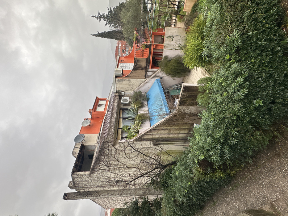 The image displays a multi-level residential building complex situated in Budva, Montenegro, under an overcast sky. The main structure features a concrete facade, extensively covered on the left side by dormant climbing vines, and distinct sections painted a bright orange. Two black satellite dishes are mounted on the upper orange wall. A bare-branched deciduous tree is positioned in the foreground to the left of the building.
The mid-section of the building includes a window, an exterior air conditioning unit, and a planter containing several large, succulent-type plants. Below this, a light blue retractable awning extends over a lower patio area. Within this covered space, a silver step ladder and two green plastic chairs are visible. A paved pathway runs horizontally across the lower-left foreground, separated from the building's grounds by a dense, dark green hedge.
To the right of the central structure, another orange-painted section features a balcony with a black metal railing and multiple potted plants. The surrounding grounds incorporate various green shrubs and several evergreen trees. In the background, distant hills and additional residential buildings with red roofs are visible beneath a uniformly cloudy sky. No individuals are present in the scene.