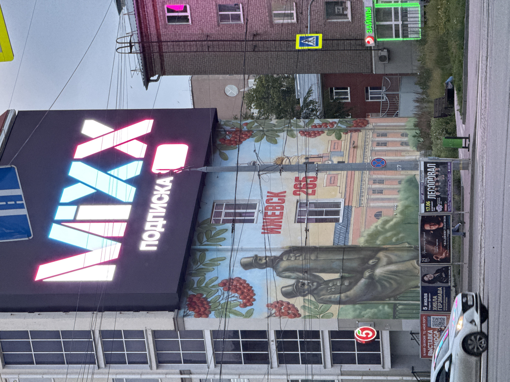 This is a medium shot of a street scene in Izhevsk, Russia. The most prominent feature is a large, illuminated billboard displaying the word "MIXX" in large, neon-pink and blue letters, with "ПОДПИСКА" in smaller white letters below. To the left of the billboard, a multi-story building with numerous windows is visible. A mural painted on the side of a building depicts two men in historical attire, along with red rowan berries and green leaves. Below the mural, several posters are displayed, including one advertising an exhibition and another promoting an event with a performer named Vladimir Glazunov. A white car is driving past on the street in the foreground. To the right, a brick building with several windows has a green neon sign that reads "ЗАЙМЫ," indicating a financial service. A yellow pedestrian crossing sign is also visible on the right side of the street. The sky is overcast.