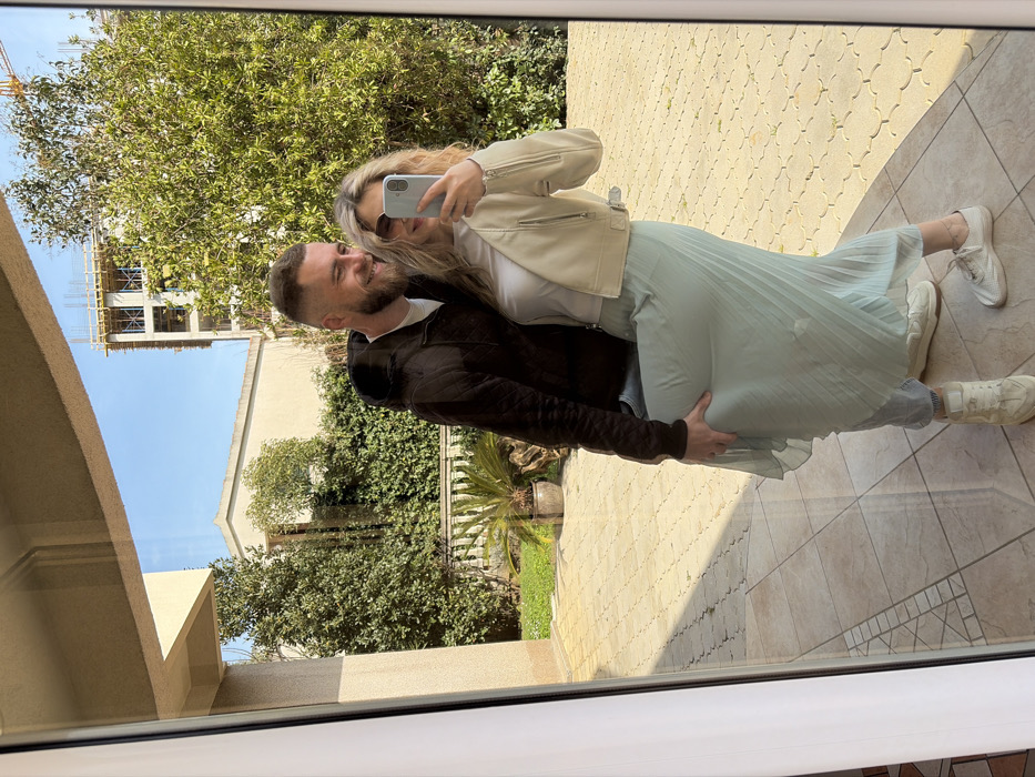 The image depicts a reflection of two individuals, a male and a female, captured in a large glass panel, likely a window or door, in Budva, Montenegro.
The female, positioned slightly in front, is on the right side of the reflection. She has blonde, wavy hair and wears sunglasses with dark lenses. Her attire consists of a white top, a light beige/cream cropped faux leather jacket with zippered details, a long, pleated, light teal skirt, and white sneakers. She holds a light-colored smartphone in her left hand, raised to eye level, obscuring part of her face as she appears to be taking a selfie. Her right arm is positioned around the male's waist.
The male is behind the female, on the left side of the reflection. He has short dark hair, a short beard, and is smiling broadly, looking towards the smartphone camera. He is wearing a dark brown or black quilted hooded jacket, a white t-shirt, and light-colored trousers, partially visible beneath the female's skirt. His left hand rests on the female's upper left thigh, lightly lifting the fabric of her skirt. His right arm is around her back.
The background, visible through and around the reflection, shows an outdoor setting under a clear blue sky, indicating bright daylight. The immediate ground outside the glass is paved with light-colored, interlocking concrete pavers. Further back, lush green vegetation, including hedges and trees, is prominent. A stone balustrade and a large potted plant (possibly a cycad) are visible. In the distance, a finished building with light-colored walls stands, and to its right, a multi-story building under construction is visible, characterized by exposed concrete framework, scaffolding, and a faint yellow crane jib overhead.