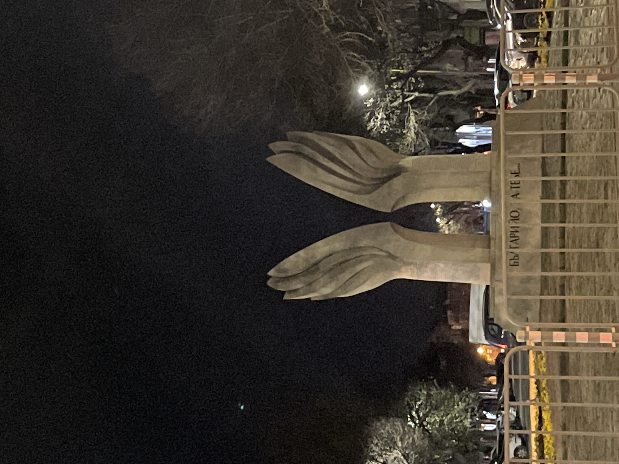 At night, the image depicts a monument in Sofia, Bulgaria, featuring two stylized, opposed hands, likely made of stone, against a dark sky. The monument is partially obstructed by a metal fence or barrier in the foreground. Text in Bulgarian is visible on a portion of the monument, reading “БЪЛГАРИ Ю. А ТЕ БЕ…”. Behind the monument, trees and streetlights are visible, and what appear to be parked cars line the street. The overall environment suggests an urban setting.