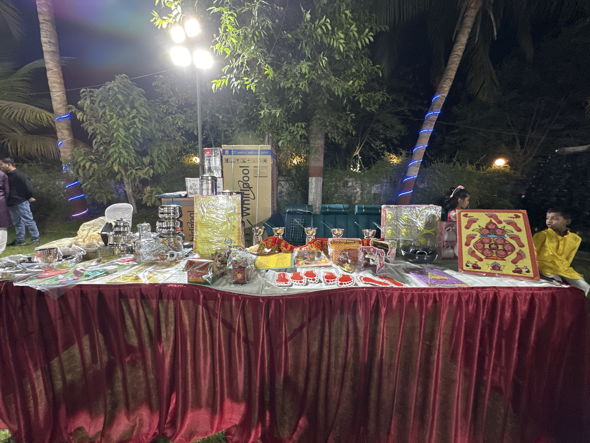 Captured at night in what appears to be Shahapur, India, the image centers on a table draped with a red, textured tablecloth. The table is laden with an assortment of items: stainless steel cookware, wrapped boxes, and decorative religious objects suggesting a ceremonial or festive occasion. Two children are positioned to the right side of the table. The background features trees, one of which is decorated with blue lights, and other individuals in the periphery, contributing to the impression of an outdoor gathering. An additional light source illuminates the scene.