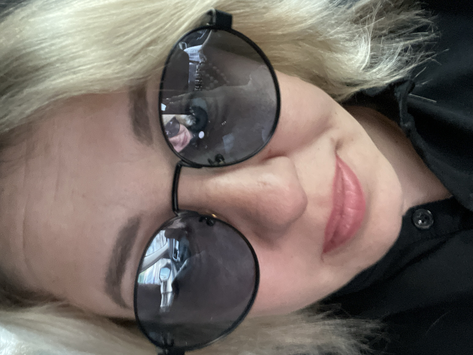 A close-up, rotated image shows a person with fair skin and blonde hair wearing round, black-rimmed sunglasses. The reflection in the sunglasses shows a car interior and cityscape suggesting the photo was taken inside a vehicle. A black buttoned shirt or jacket is also visible. It is impossible to verify the location as Chaikovsky, Russia. The image only captures a portion of the person's face. The person is slightly smiling with pink lips.