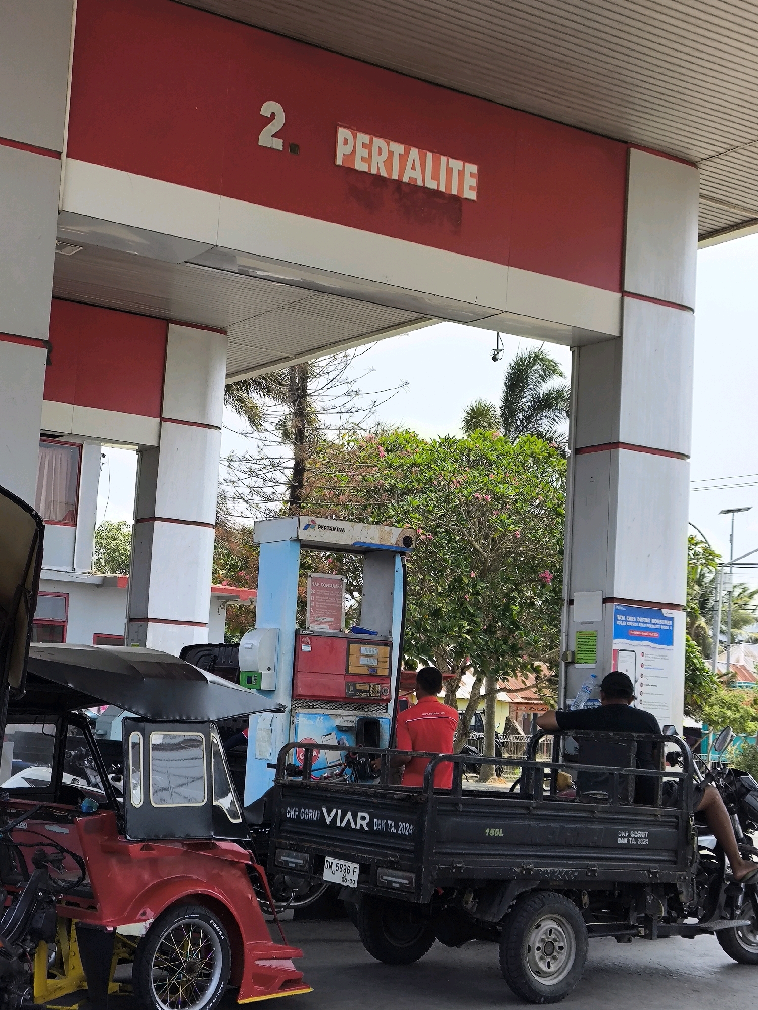 The image shows a Pertamina gas station in Unknown, Indonesia. A red and white sign above reads "2. PERTALITE". In the foreground, a red auto-rickshaw with a black canopy is parked. Behind it, a black utility vehicle with a wooden bed is present, with two men in it. One man in a red shirt is standing near a Pertamina fuel pump, while another man in a black t-shirt is seated in the utility vehicle. The fuel pump is blue and red, with multiple display screens and nozzles. Trees and some buildings are visible in the background. The scene appears to be during daylight hours, with a clear sky.