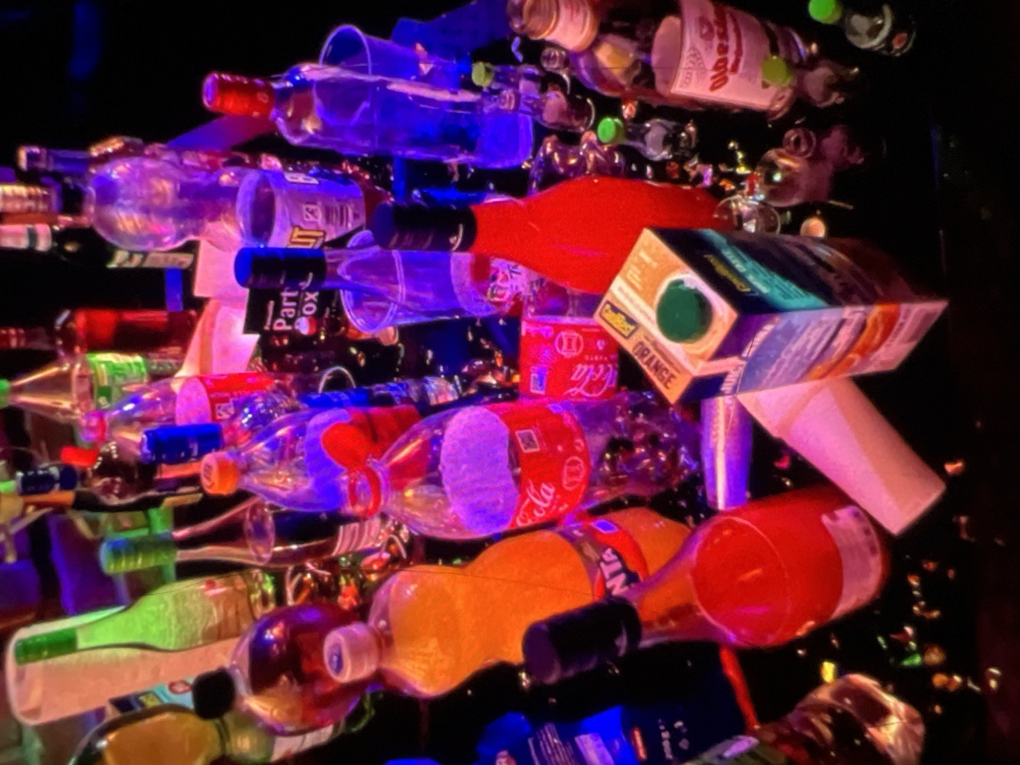The image depicts a dimly lit, cluttered table surface, possibly in a nightclub or party setting in Lagos, Nigeria. The scene is dominated by numerous bottles of alcoholic beverages and soft drinks, many of which are partially or fully consumed. Several plastic cups, some filled with liquids and others empty, are scattered among the bottles. Confetti or small pieces of paper litter the surface. The lighting is artificial and colorful, with prominent purple and pink hues, suggesting a vibrant nightlife atmosphere. The arrangement of the items suggests a gathering or celebration where drinks have been freely served and consumed. There are no people clearly visible in the image.
